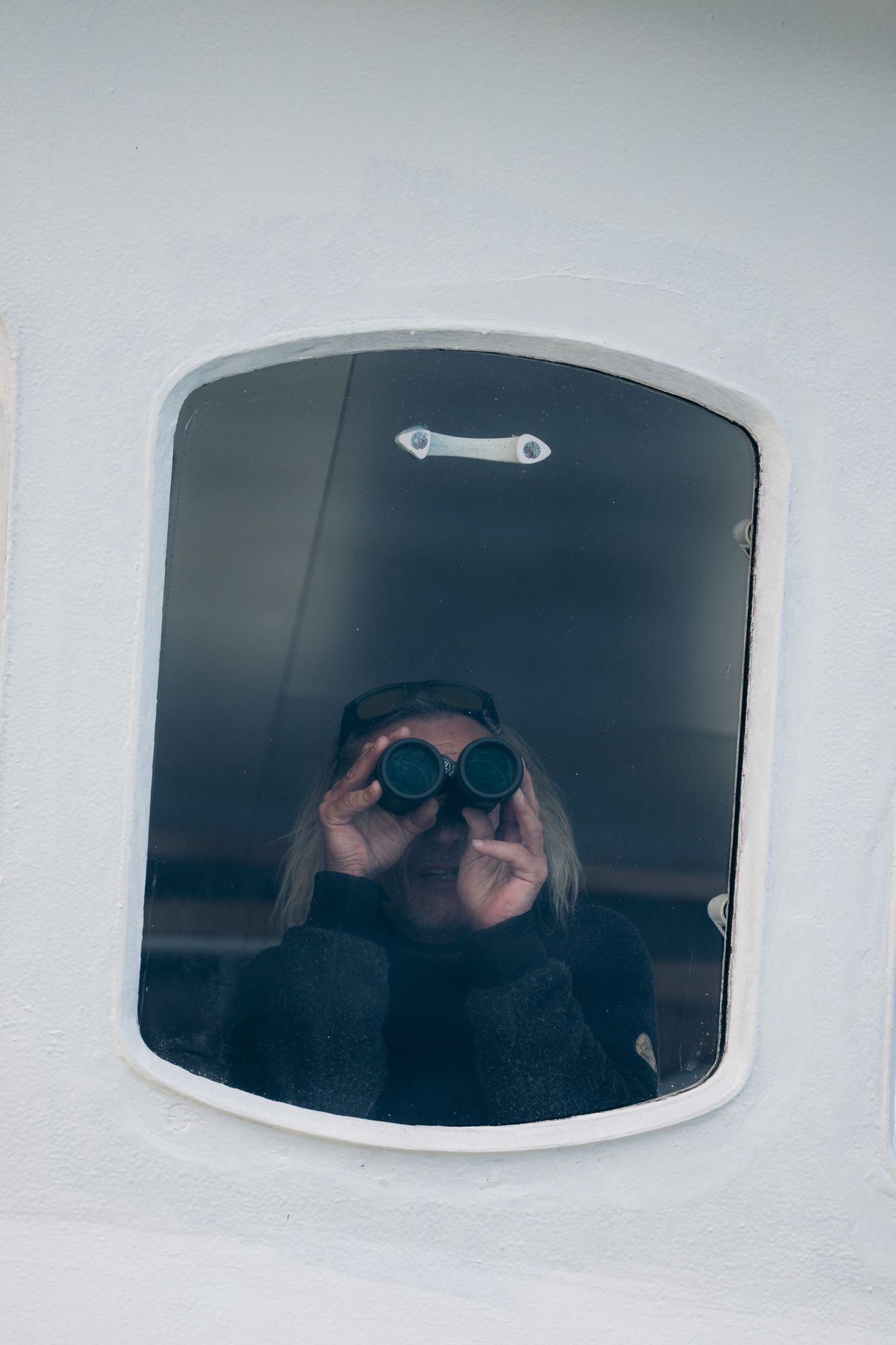 Visage d'un passager aux jumelles cadré dans un hublot de la coque blanche