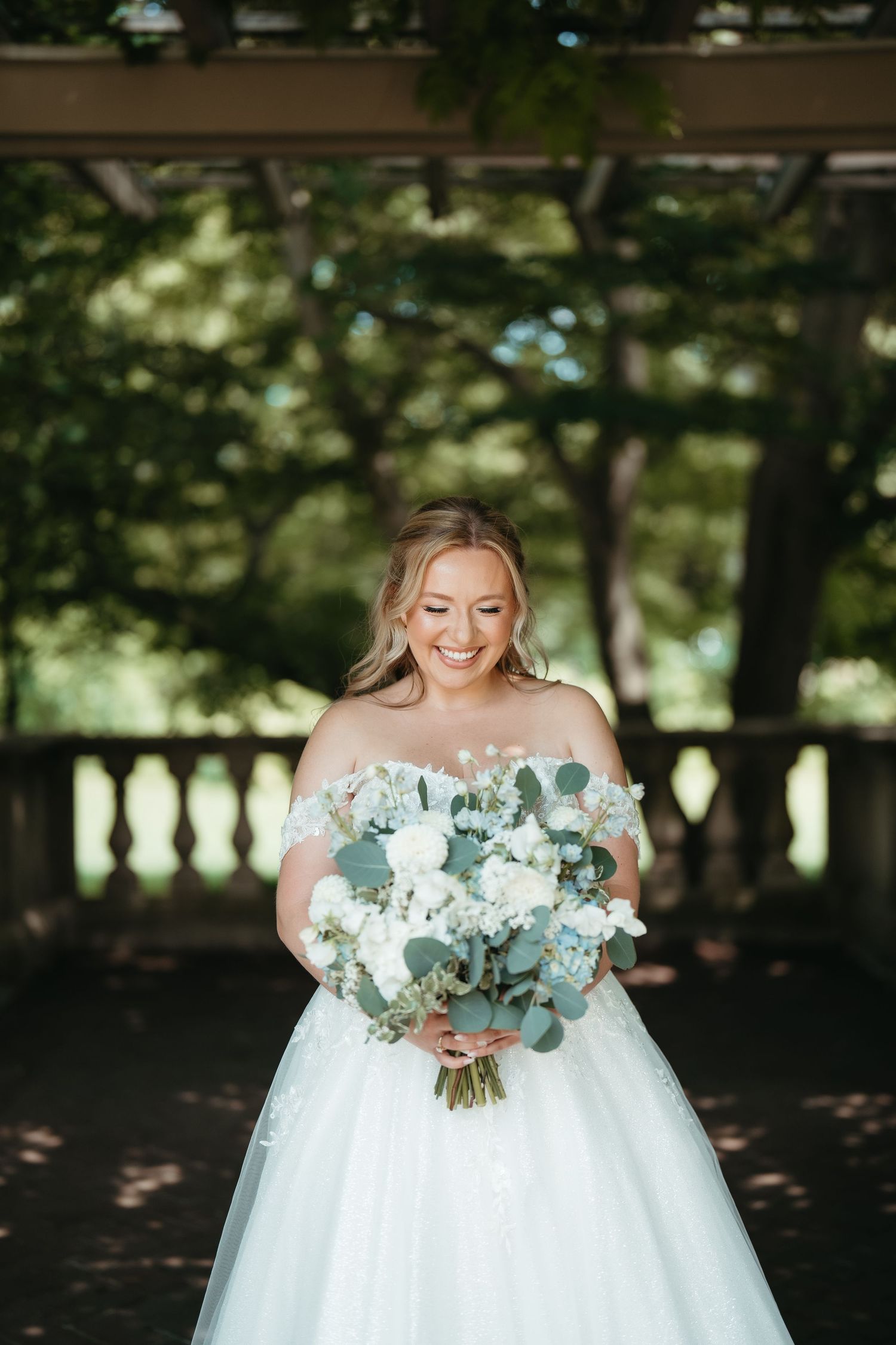 Smiling bride in off-shoulder white ball gown holding lush bouquet of white and blue flowers outdoors.