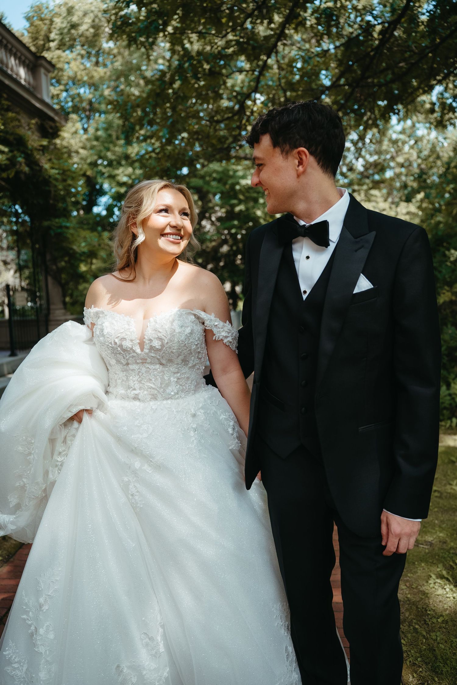 A bride in a lace off-shoulder gown and groom in a black tuxedo hold hands and smile at each other outdoors.