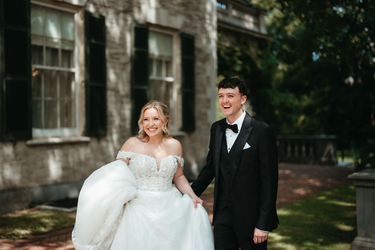 Bride in white ballgown and groom in black tuxedo laughing while holding hands outside historic building.