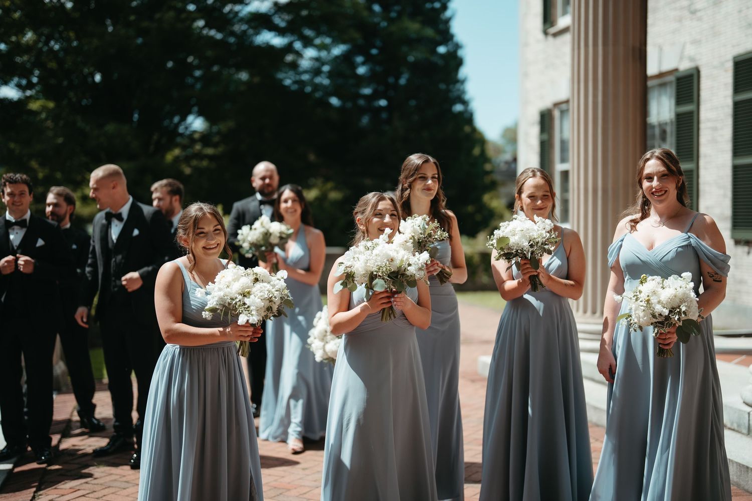 Bridesmaids in dusty blue gowns holding white bouquets walk outdoors near a columned building on a sunny wedding day.