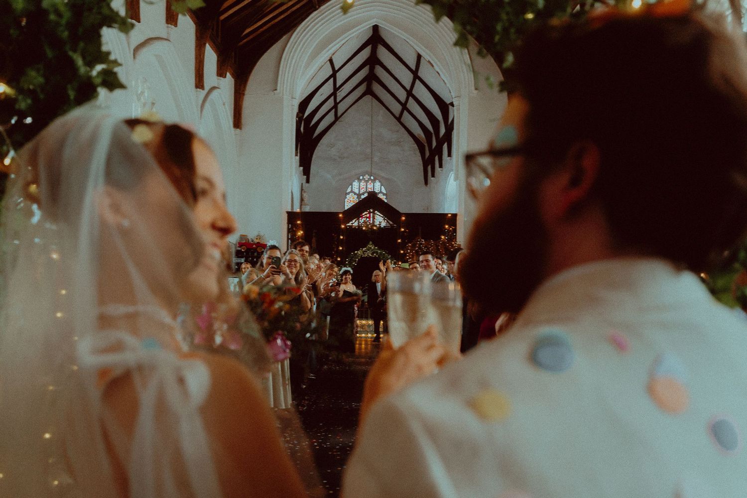 Bride and groom holding champagne glasses in a church wedding ceremony with colorful confetti.