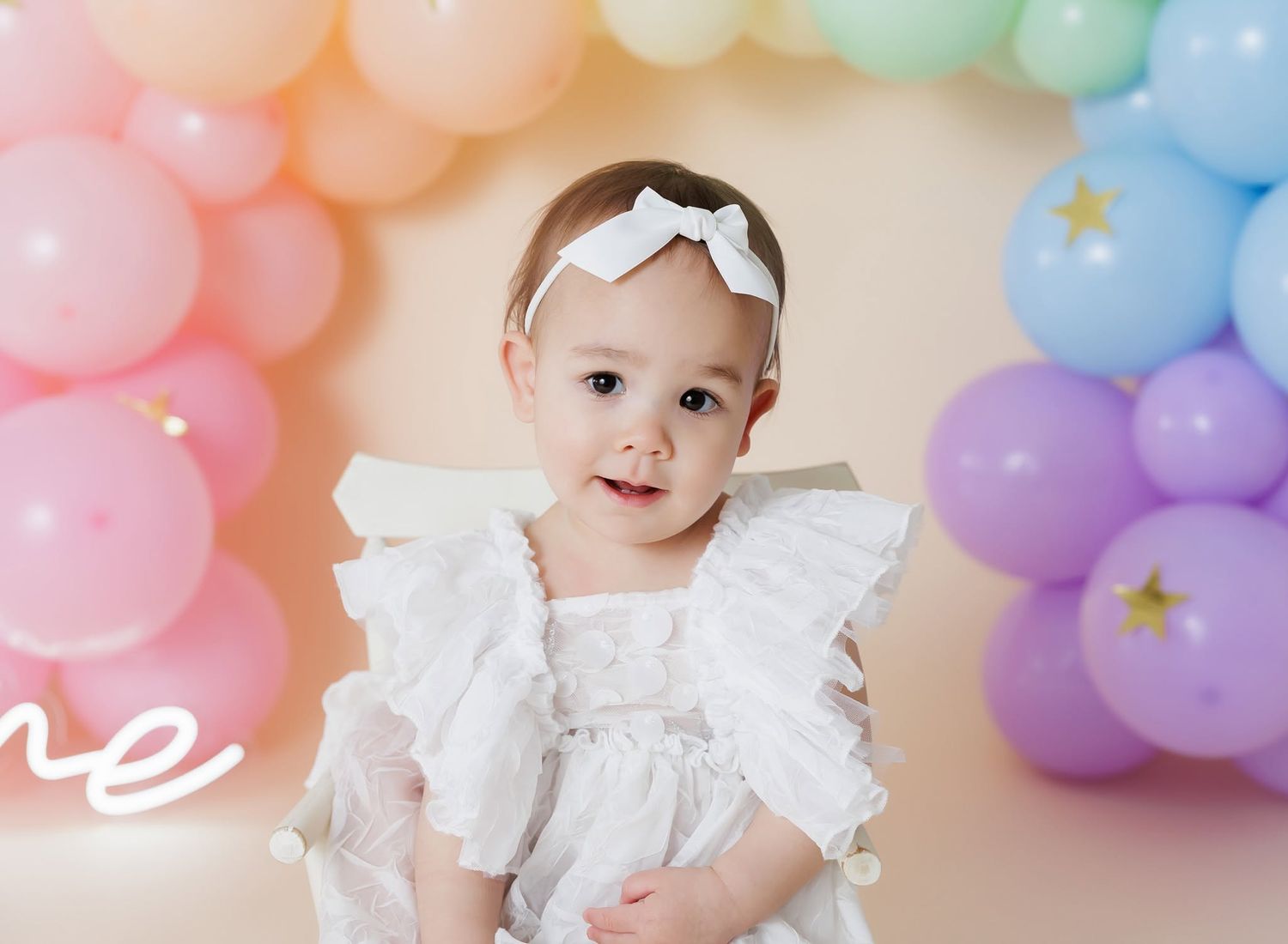 Baby girl in white dress with bow headband smiles in front of colorful pastel balloon arch at first birthday party.