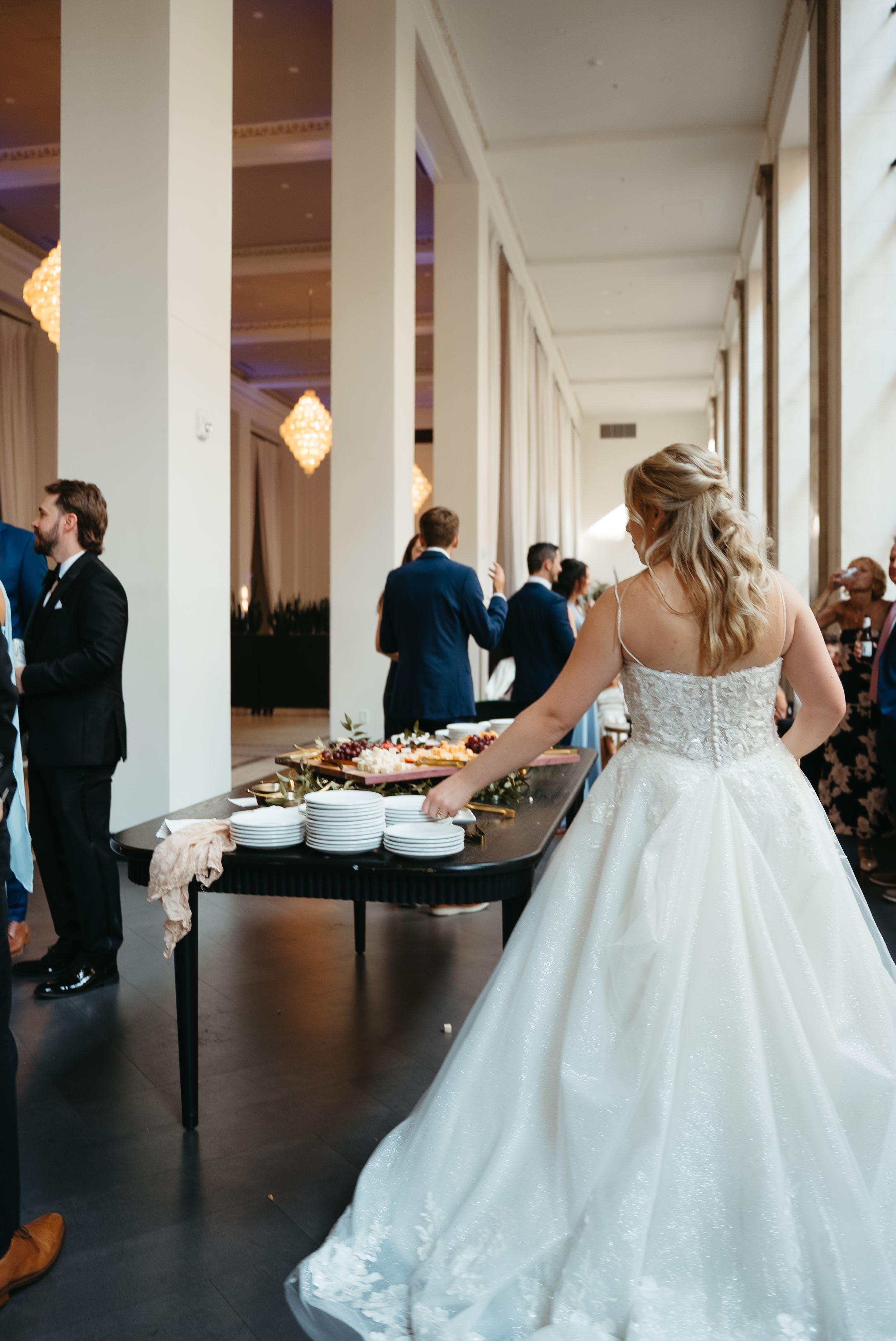 Bride in a white lace ball gown reaching for plates at a wedding reception buffet table.