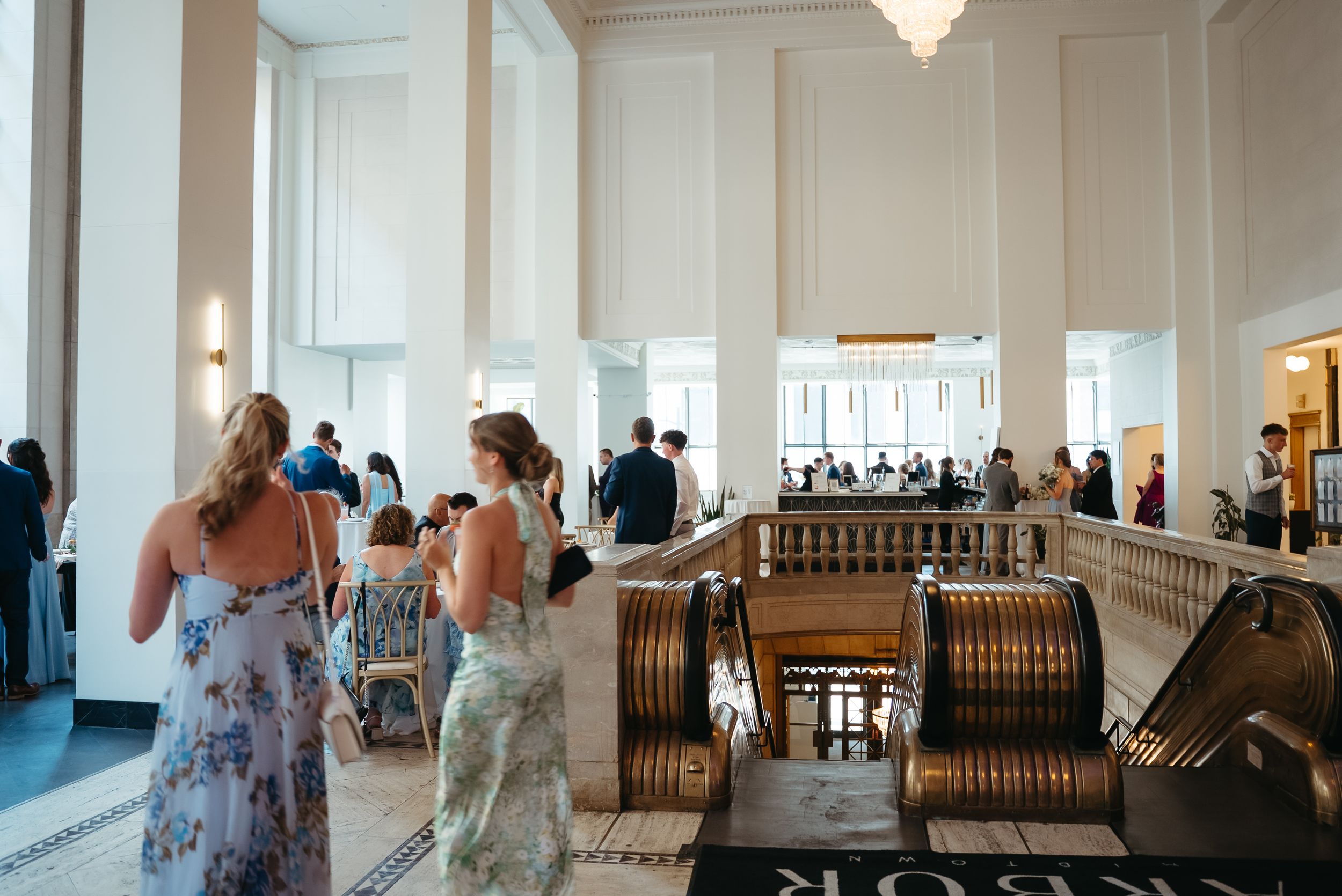 Elegant wedding reception in a grand hall with ornate columns, chandelier, and guests in floral dresses near vintage escalators.