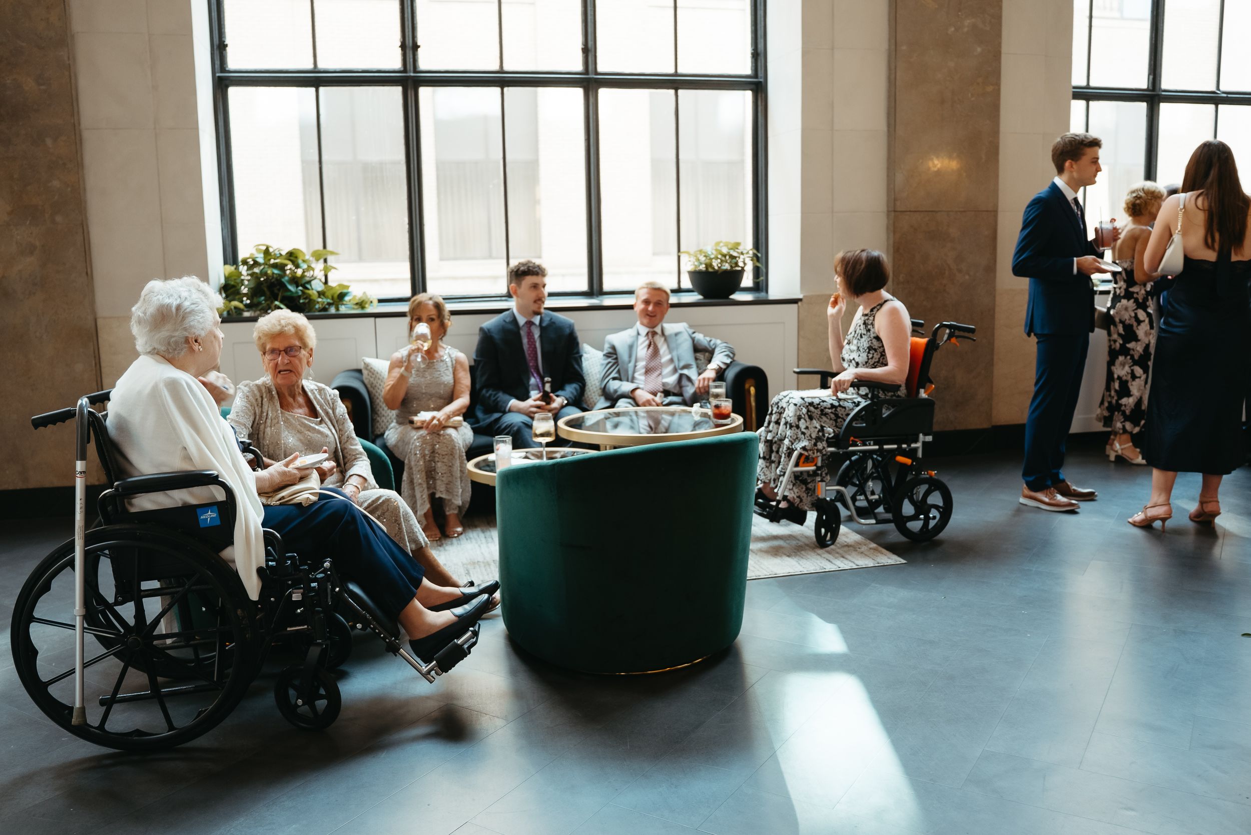 Elderly guests in wheelchairs and formally dressed attendees socialize in a bright modern venue during a special event.