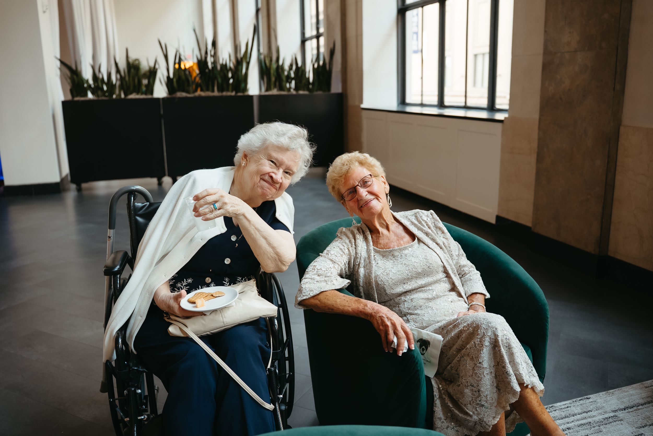 Two elderly women sharing a joyful moment together in a modern lobby, one seated in a wheelchair holding a plate of food.