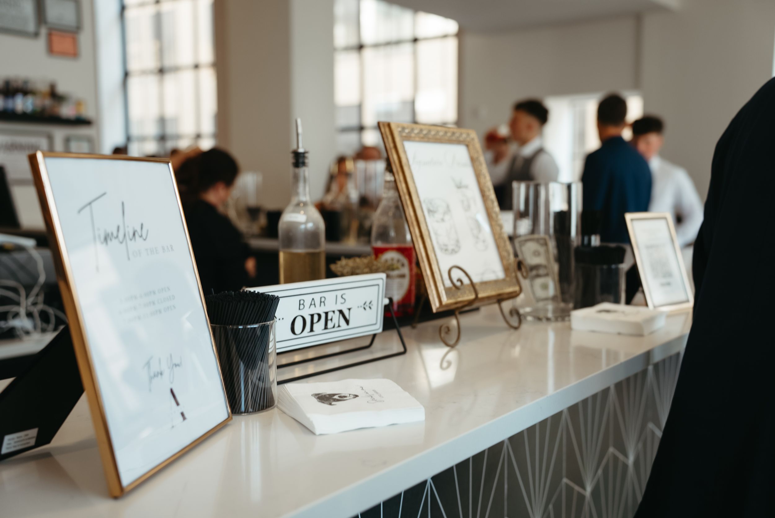 Bar counter with open sign, framed menus, and guests mingling in a bright modern venue.