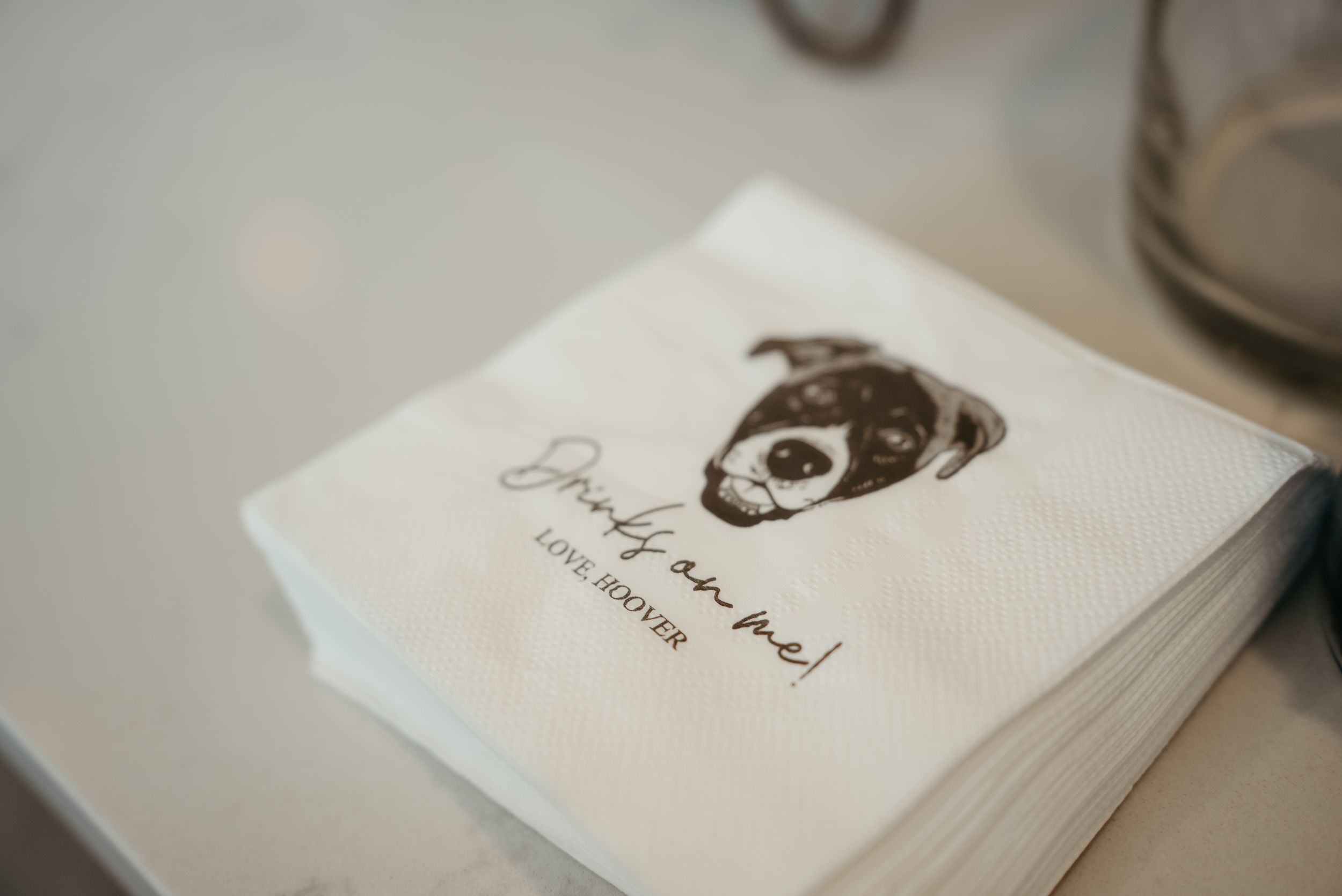 Custom white cocktail napkins with a puppy illustration and "Drinks on me! Love, Hoover" printed in brown ink.
