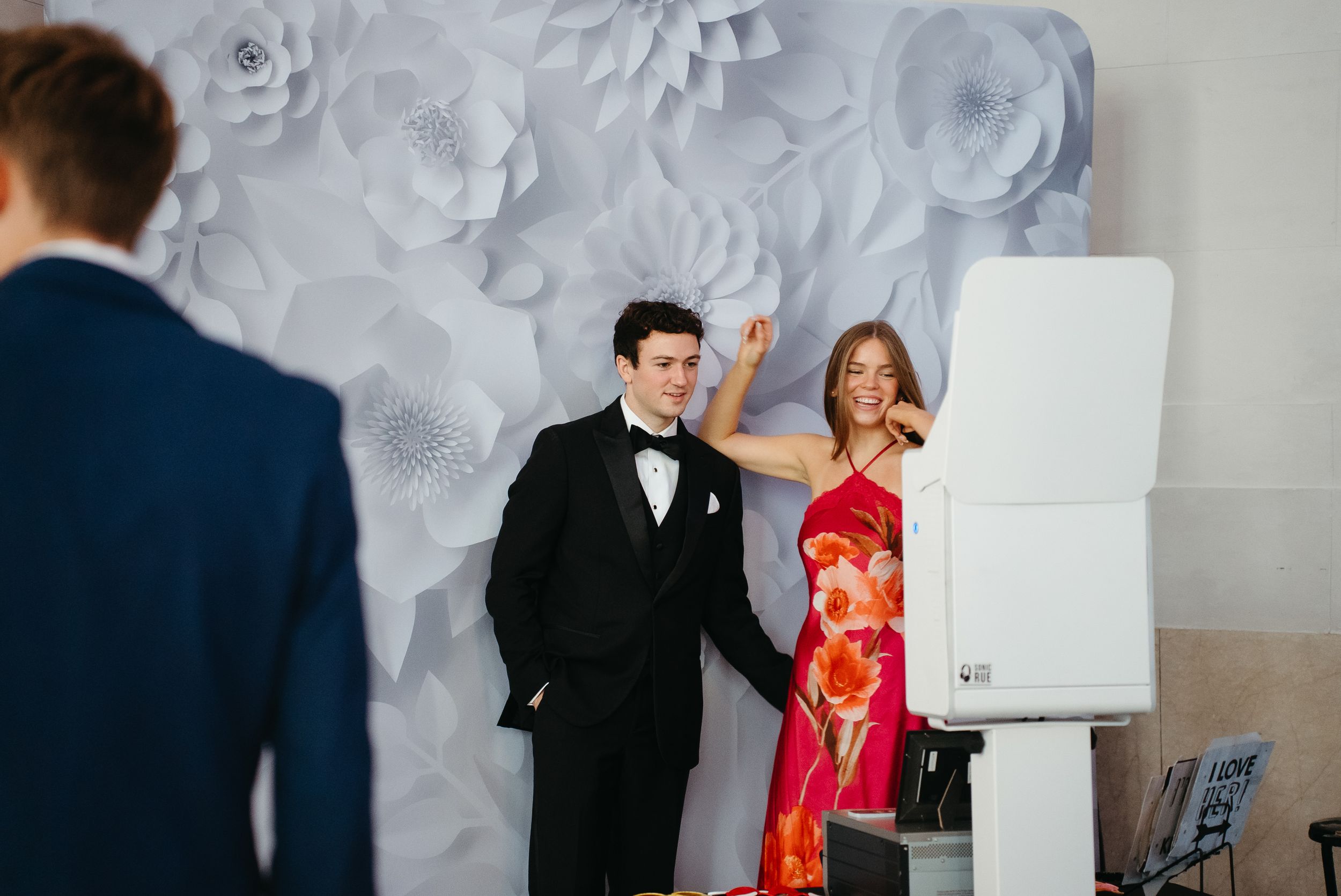 Couple posing at a wedding photo booth with white paper flower backdrop, man in tuxedo and woman in red floral dress.