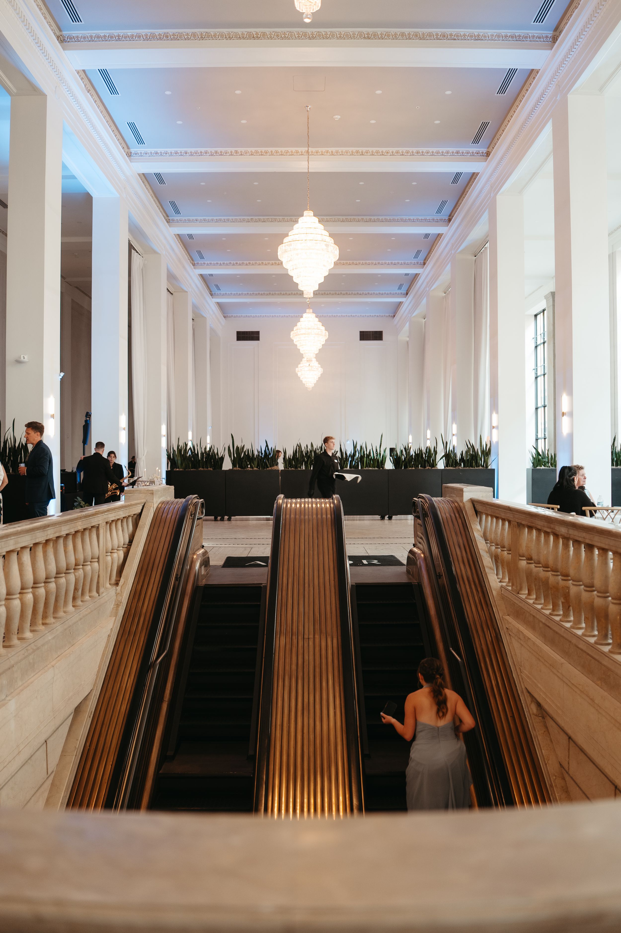 Elegant hotel lobby with escalators, white columns, coffered ceiling, and diamond chandeliers in a grand neoclassical interior.