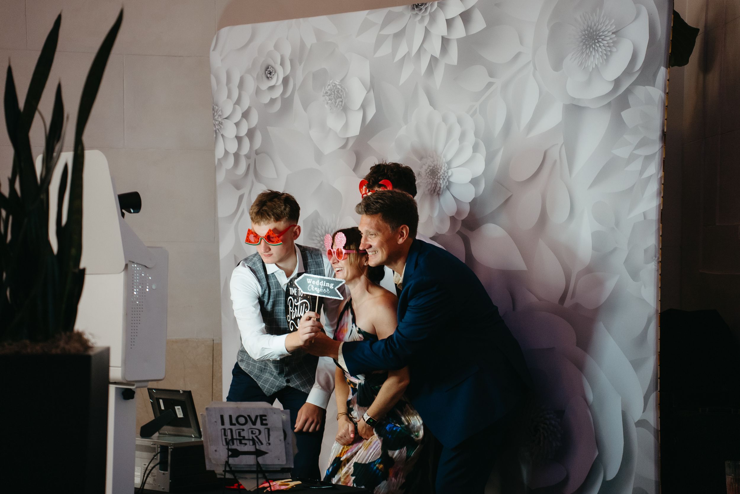 Three people pose at a photo booth with paper flower backdrop, wearing fun red glasses at a wedding reception.