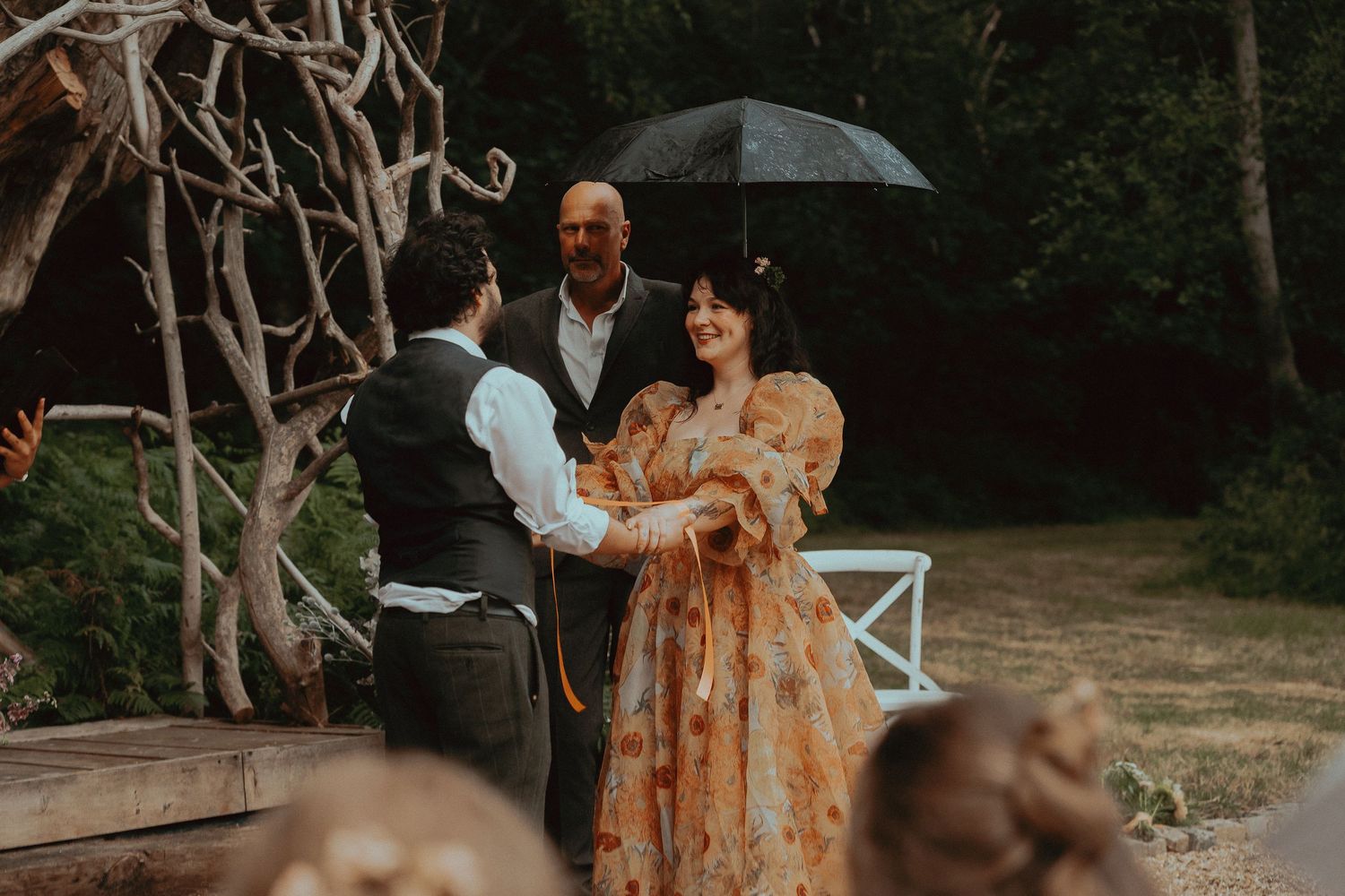 Couple exchanging vows outdoors in lush garden setting, officiant holding umbrella, bride in floral gown.
