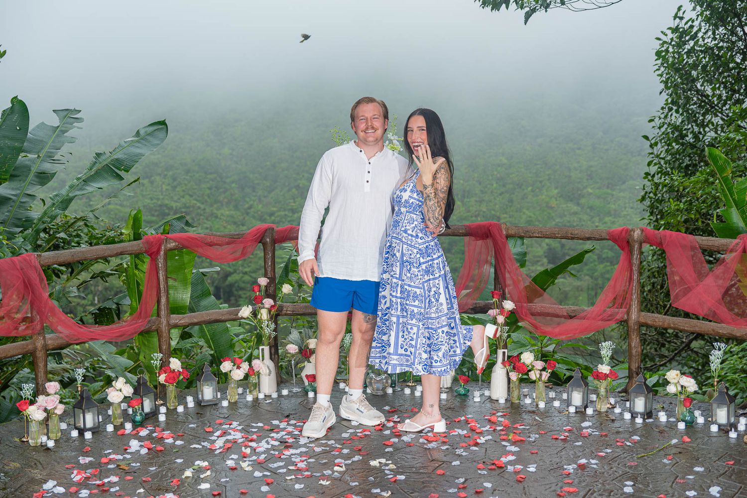 Couple posing on a romantic candle-lit overlook with rose petals and red drapes, surrounded by misty tropical jungle.