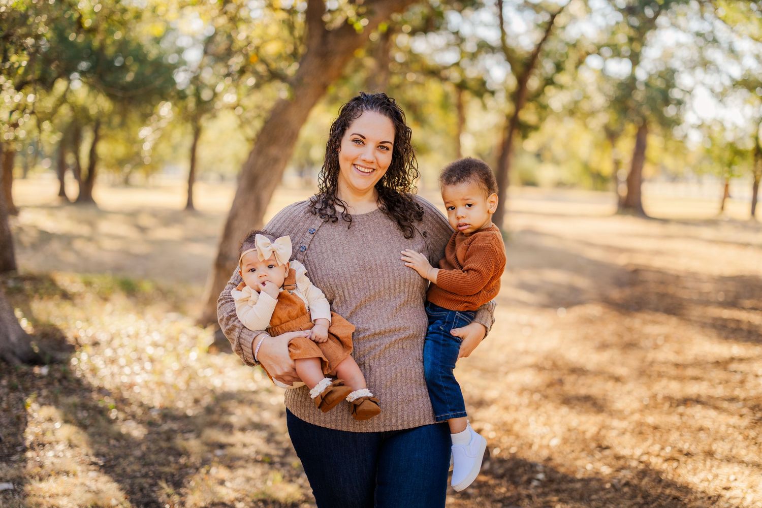 A smiling woman holds two young babies outdoors among trees in a warm autumn park setting.
