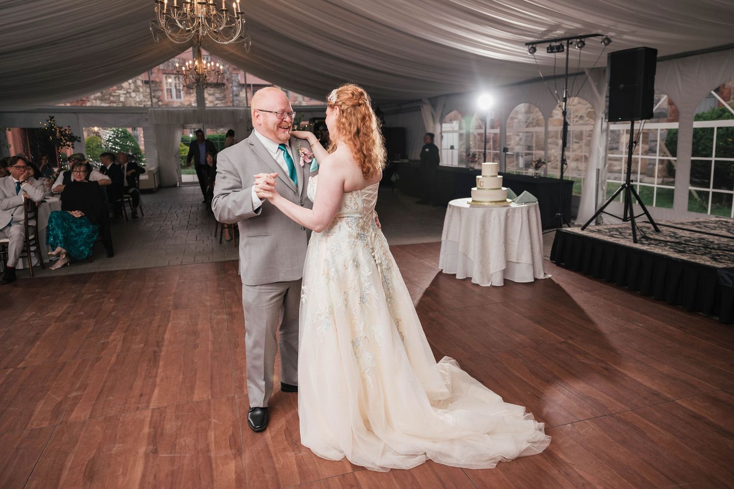 Wide view of the father-daughter dance under chandeliers, the wedding cake and DJ setup visible in the reception tent.