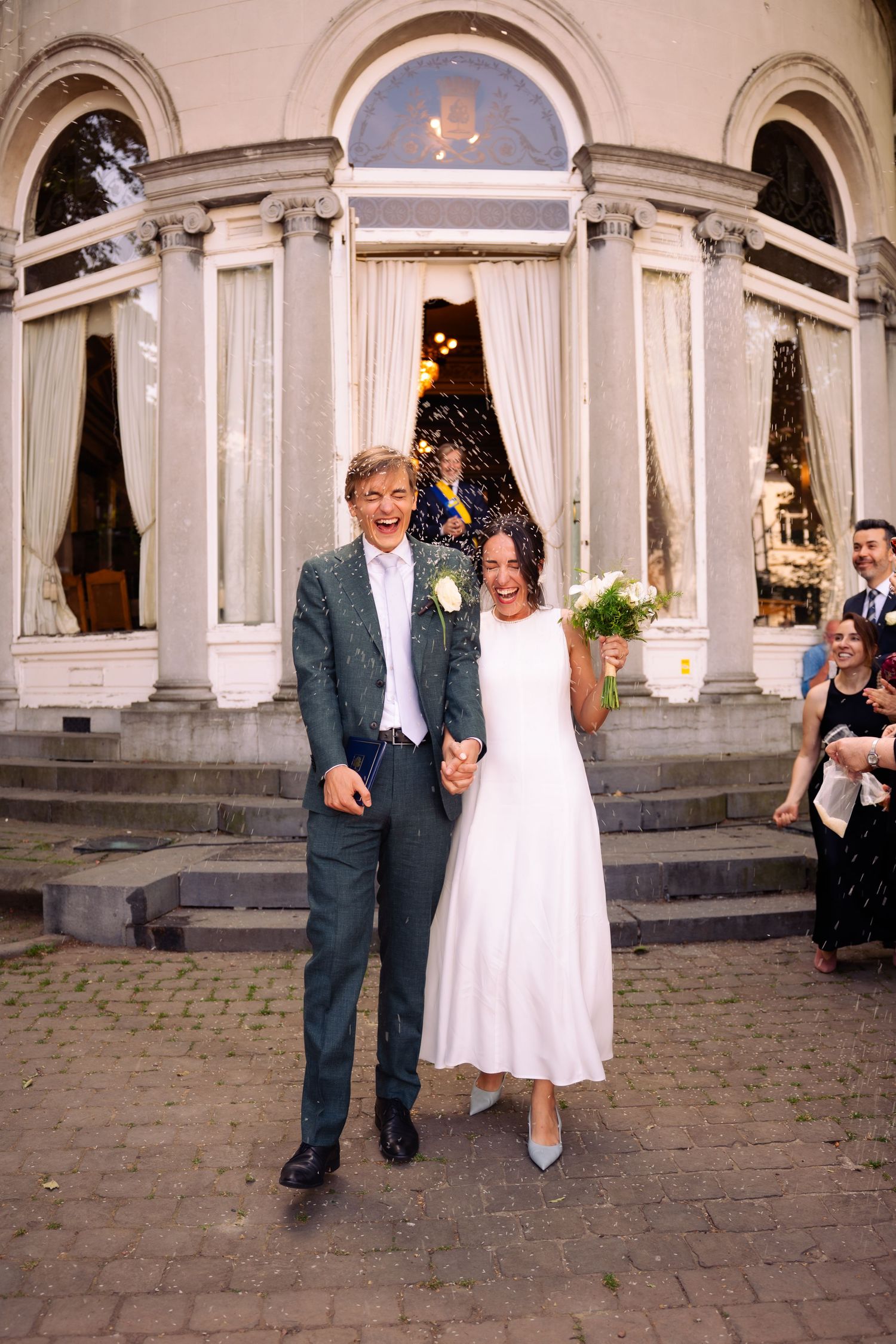 Happy newlyweds holding hands outside elegant venue, bride in white dress with bouquet, groom in gray suit, guests celebrating.