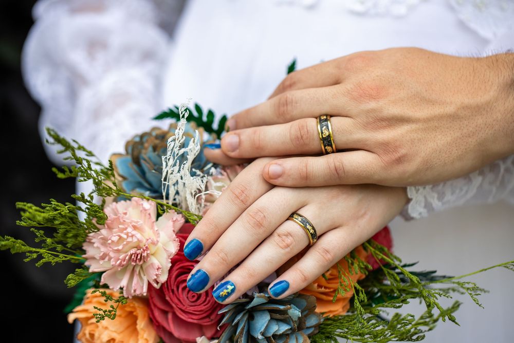 Newlywed couple displaying gold wedding rings on hands resting over a colorful bridal bouquet with blue nails.