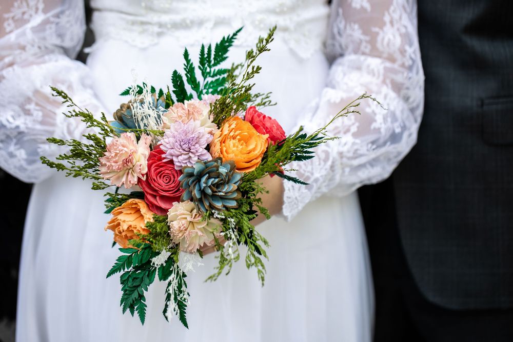 Bride and groom holding a colorful wedding bouquet with roses, dahlias, and ferns in orange, pink, and red hues.