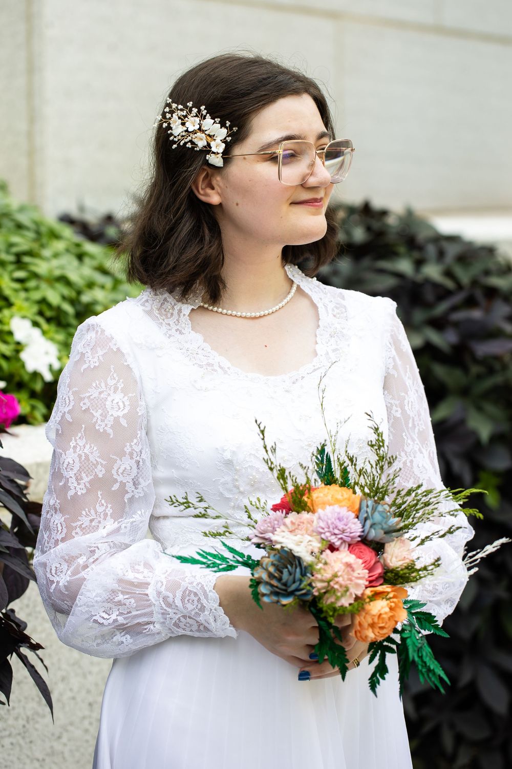 Young bride in white lace dress holding colorful bouquet with succulents, wearing pearl necklace and floral hair accessory.