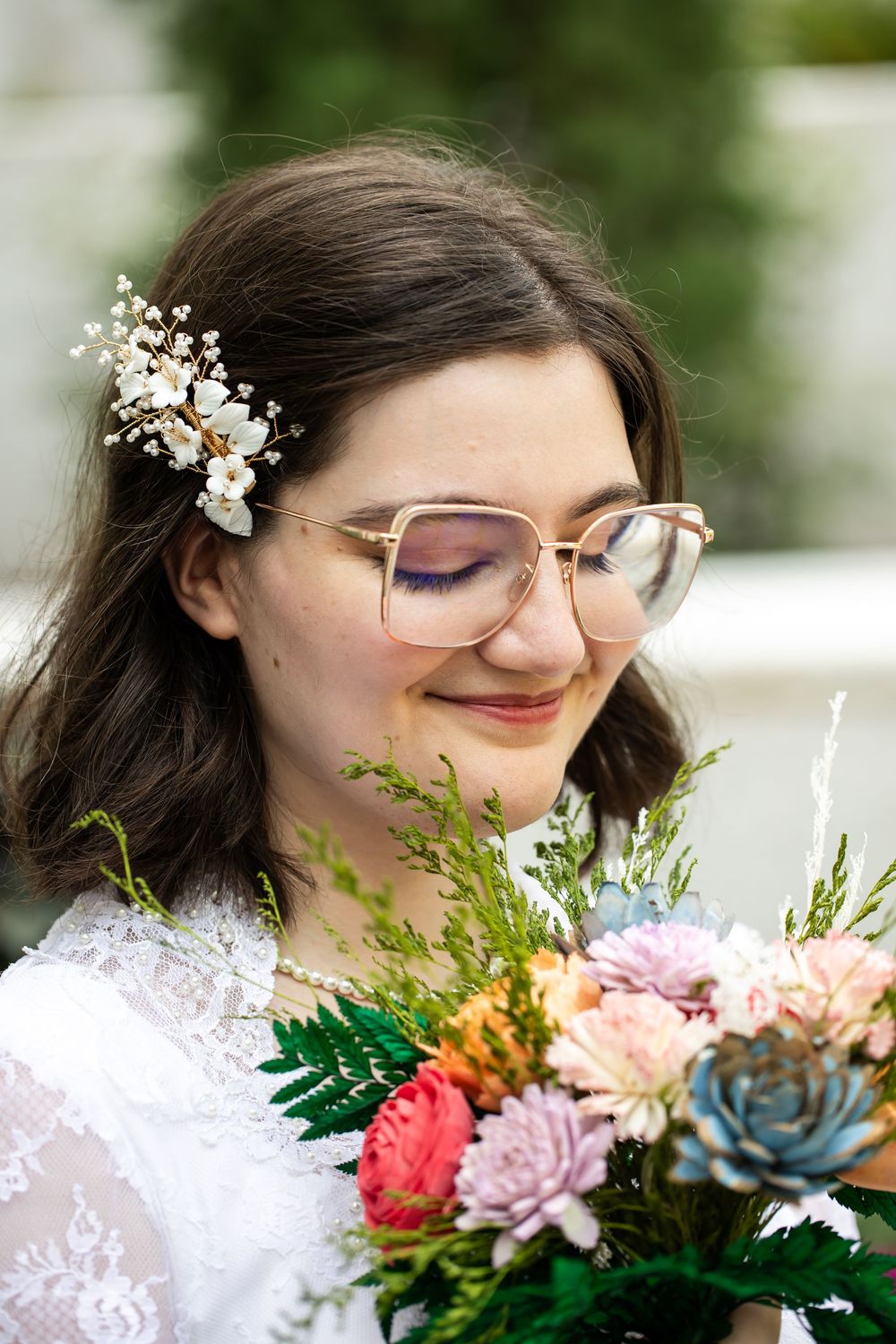 A smiling bride with glasses and floral hair accessory holds a colorful bouquet with succulents and pink flowers.