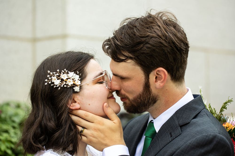 Bride and groom sharing a romantic kiss at their wedding, bride wearing floral hair accessory, groom in gray suit with green tie.