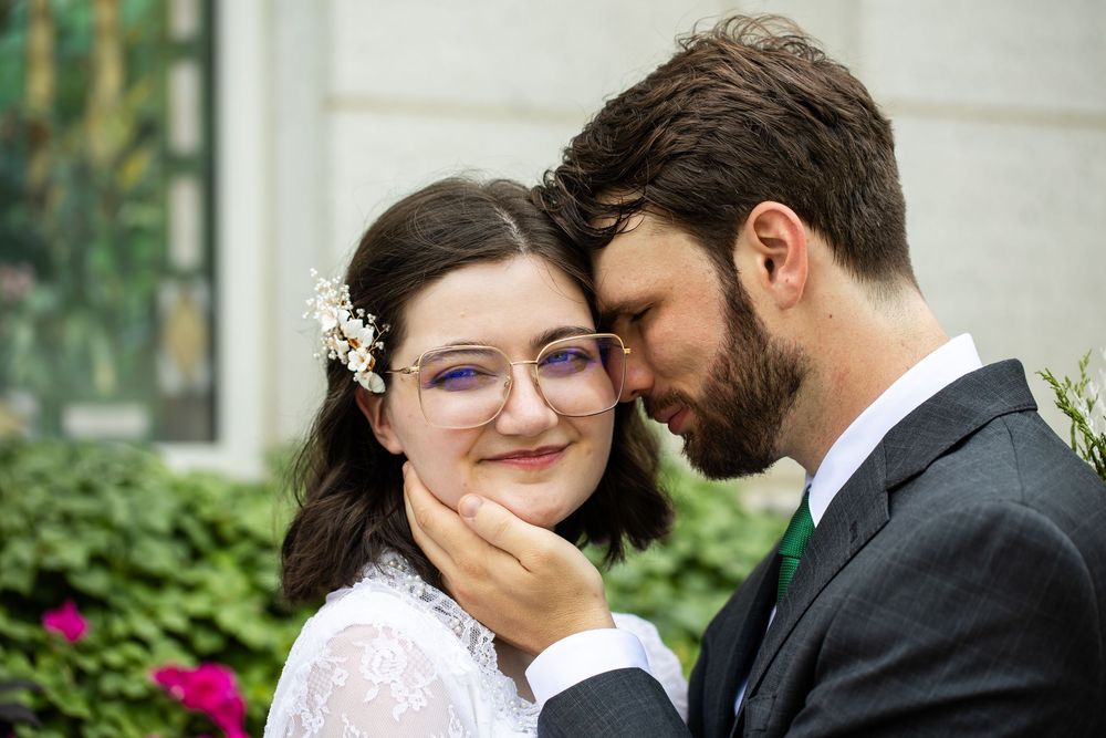 Groom tenderly holds bride's face as they touch foreheads, bride wearing glasses and floral hair accessories.