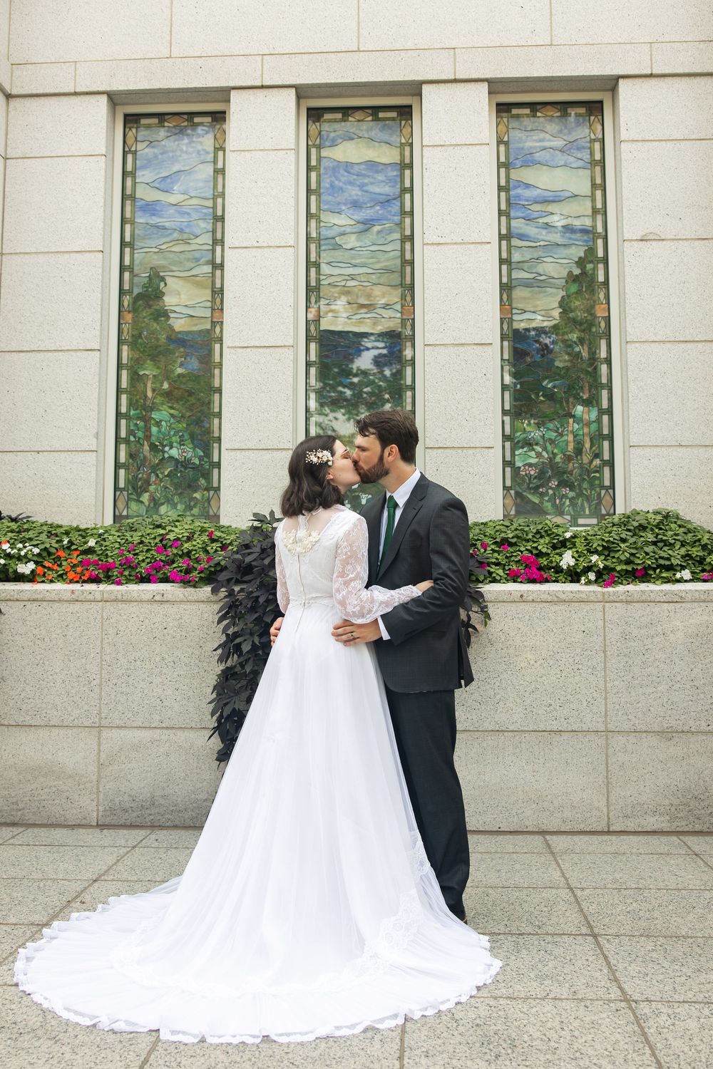 Bride and groom share a kiss outside a building with stained glass windows, bride in white lace gown with long train.