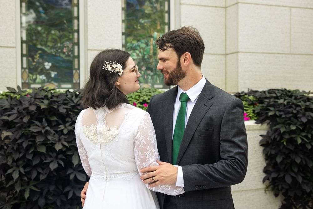 Bride and groom in wedding attire gazing at each other outdoors, man in gray suit with green tie, woman in white lace dress.
