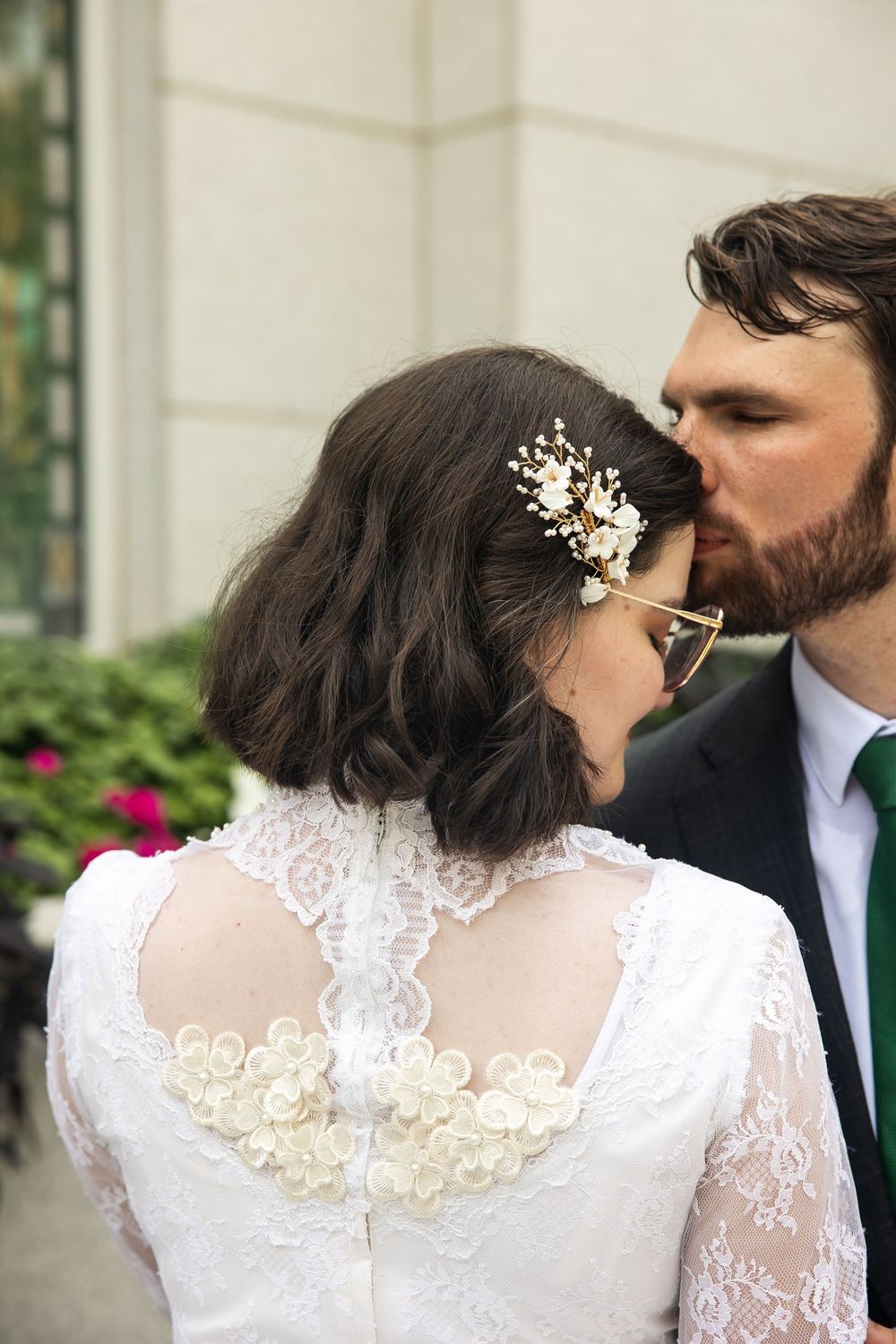 Groom kissing bride on cheek, bride wearing lace back wedding dress with floral hair accessory outdoors.