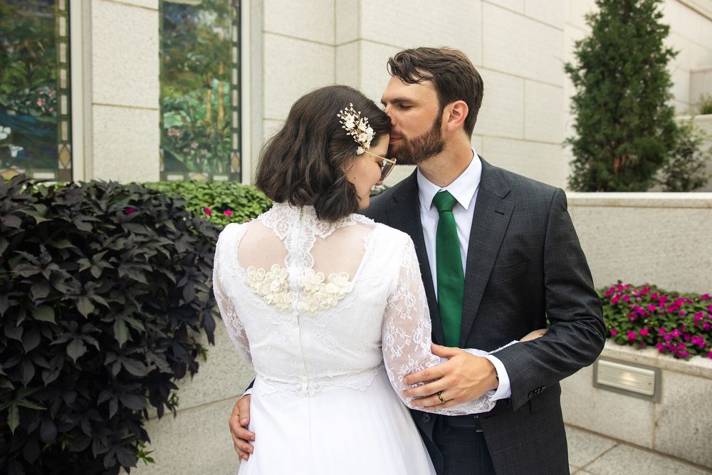 Bride and groom share a kiss outdoors, bride in lace long-sleeve gown, groom in dark suit with green tie.