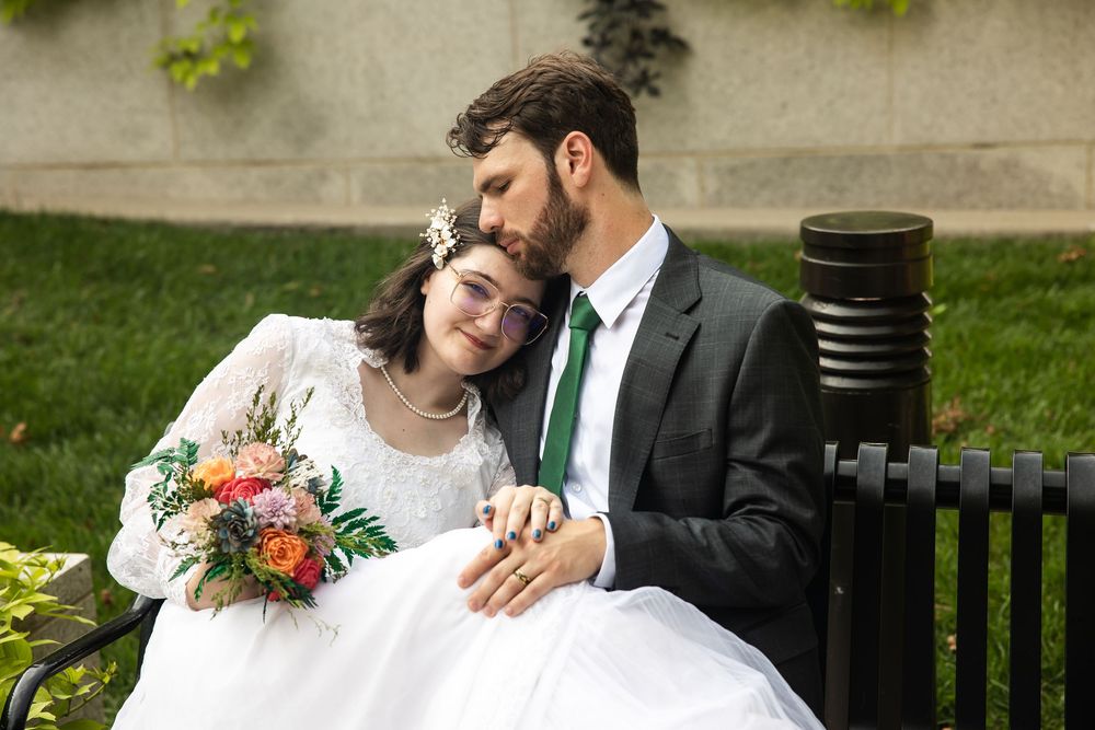 Bride and groom sitting on a bench outdoors, sharing a tender moment, bride holding a colorful bouquet in wedding attire.
