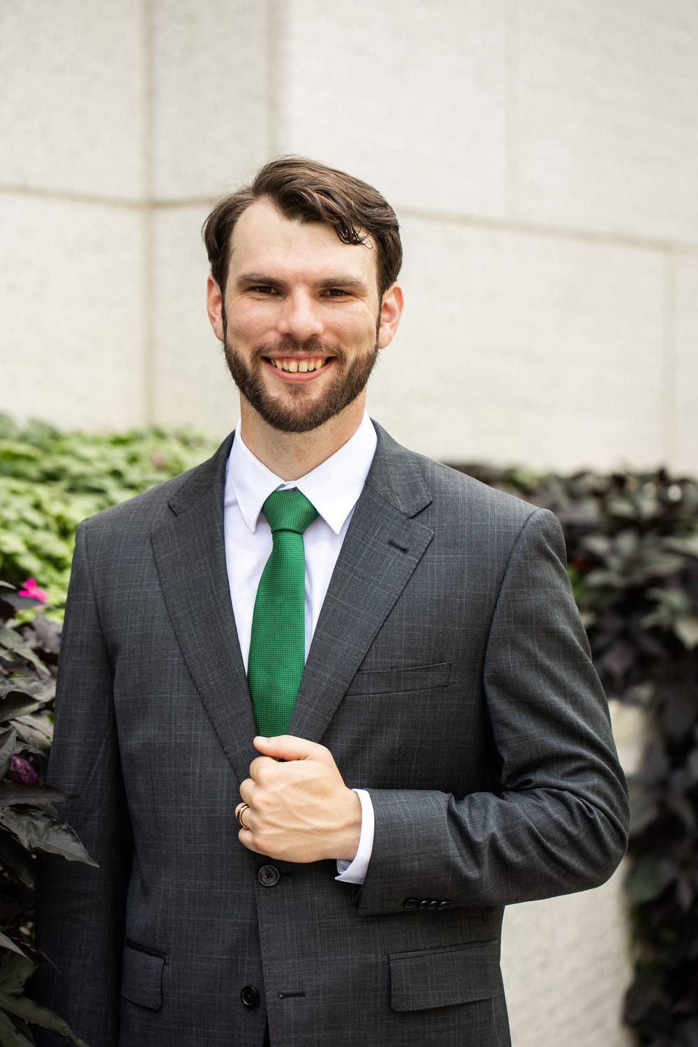 Smiling bearded man in gray suit and green tie standing outdoors near green plants and a stone wall.