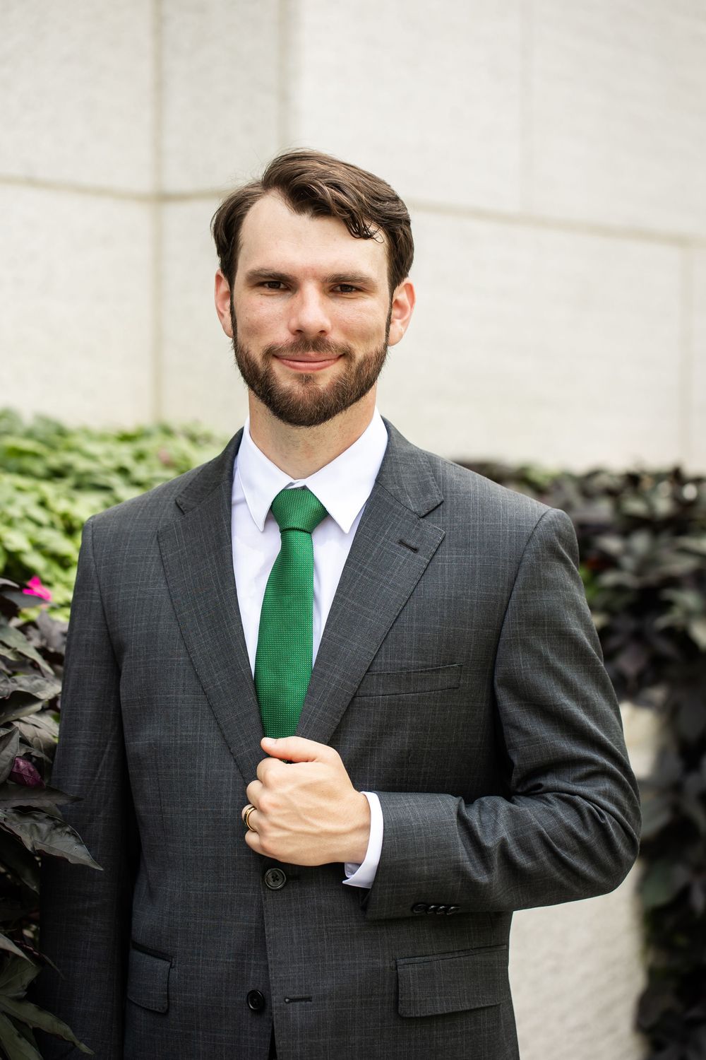 Professional man in gray suit and green tie smiling outdoors near greenery.