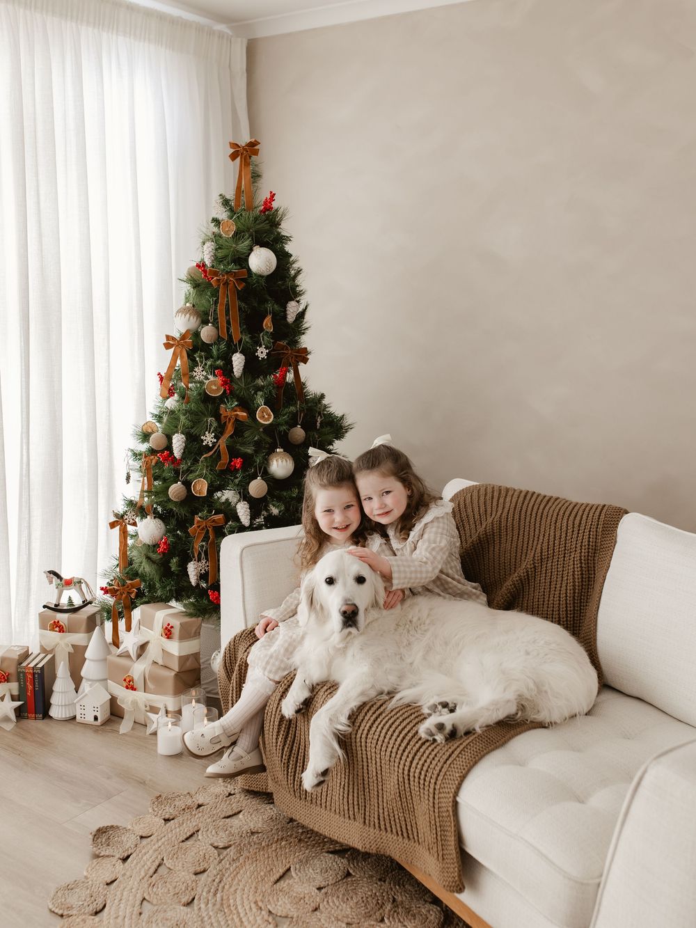 Two girls sit on a cozy sofa with a white dog beside a decorated Christmas tree with gifts.