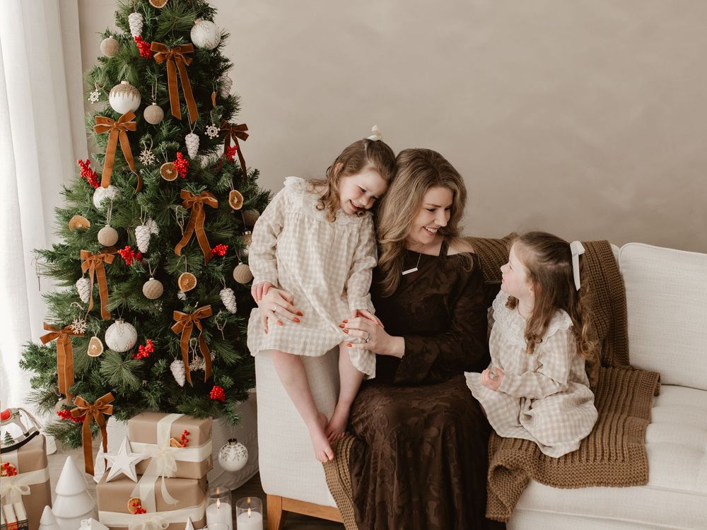 A mother and two daughters share a warm holiday moment on a white sofa beside a decorated Christmas tree with gifts.
