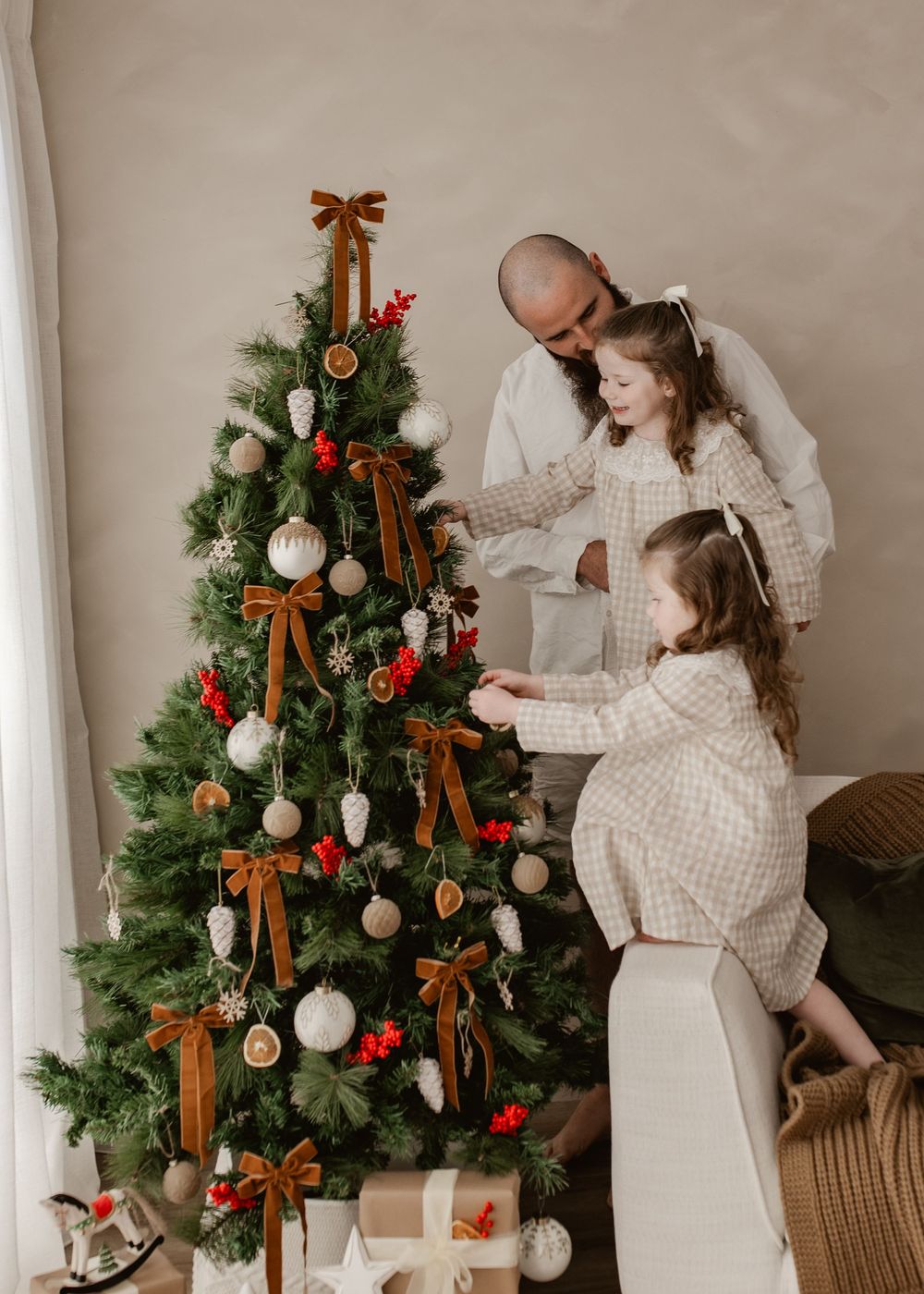 A bald father and two daughters in matching plaid dresses decorate a Christmas tree with bows and ornaments.