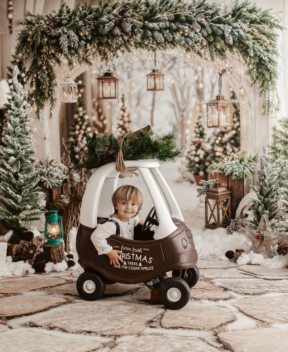 Toddler boy sits in a Christmas-themed toy car surrounded by snowy trees and lanterns in a festive holiday photo setup.