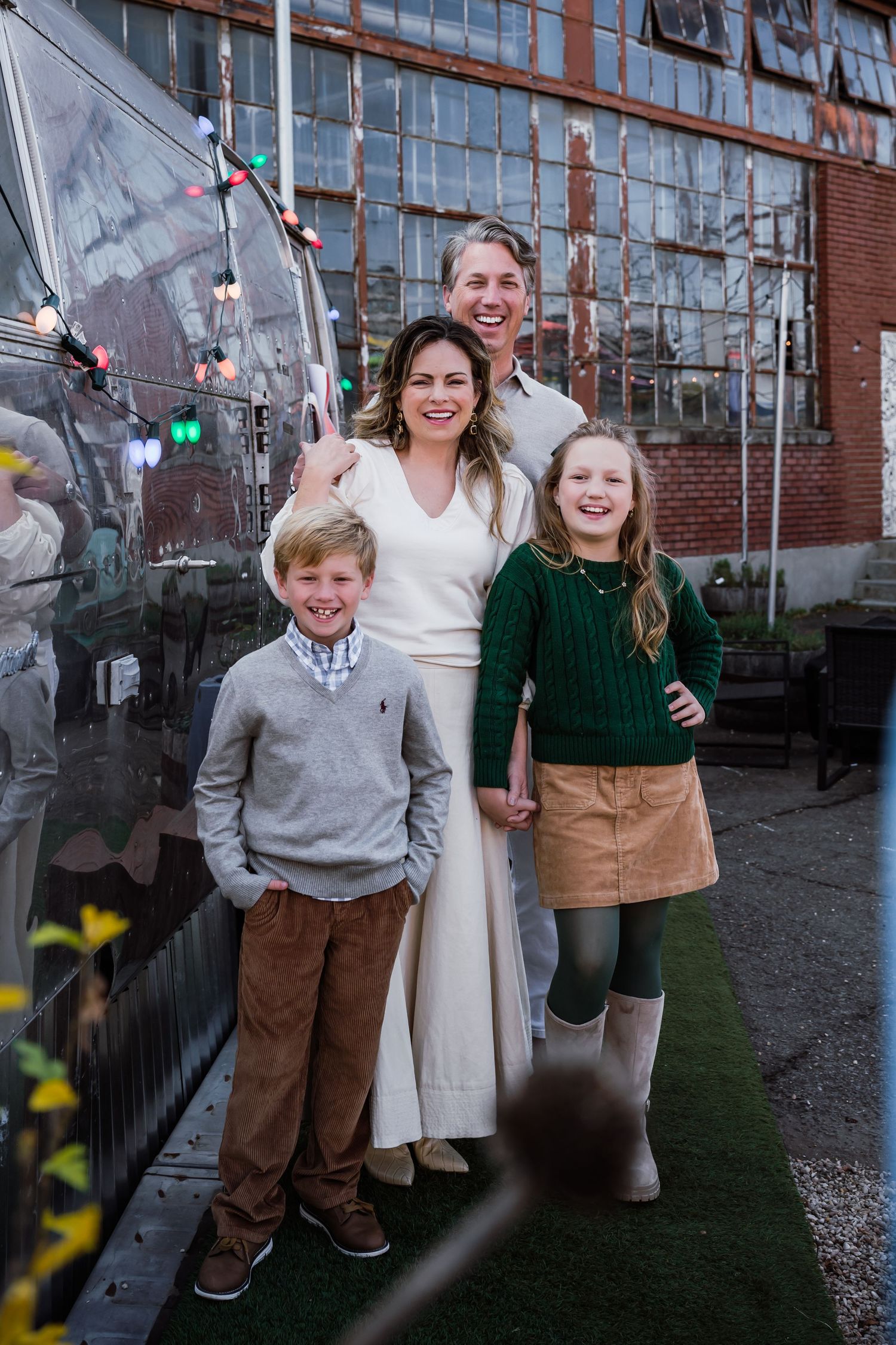 A smiling family of four poses outdoors near a silver trailer with string lights, wearing casual fall outfits.