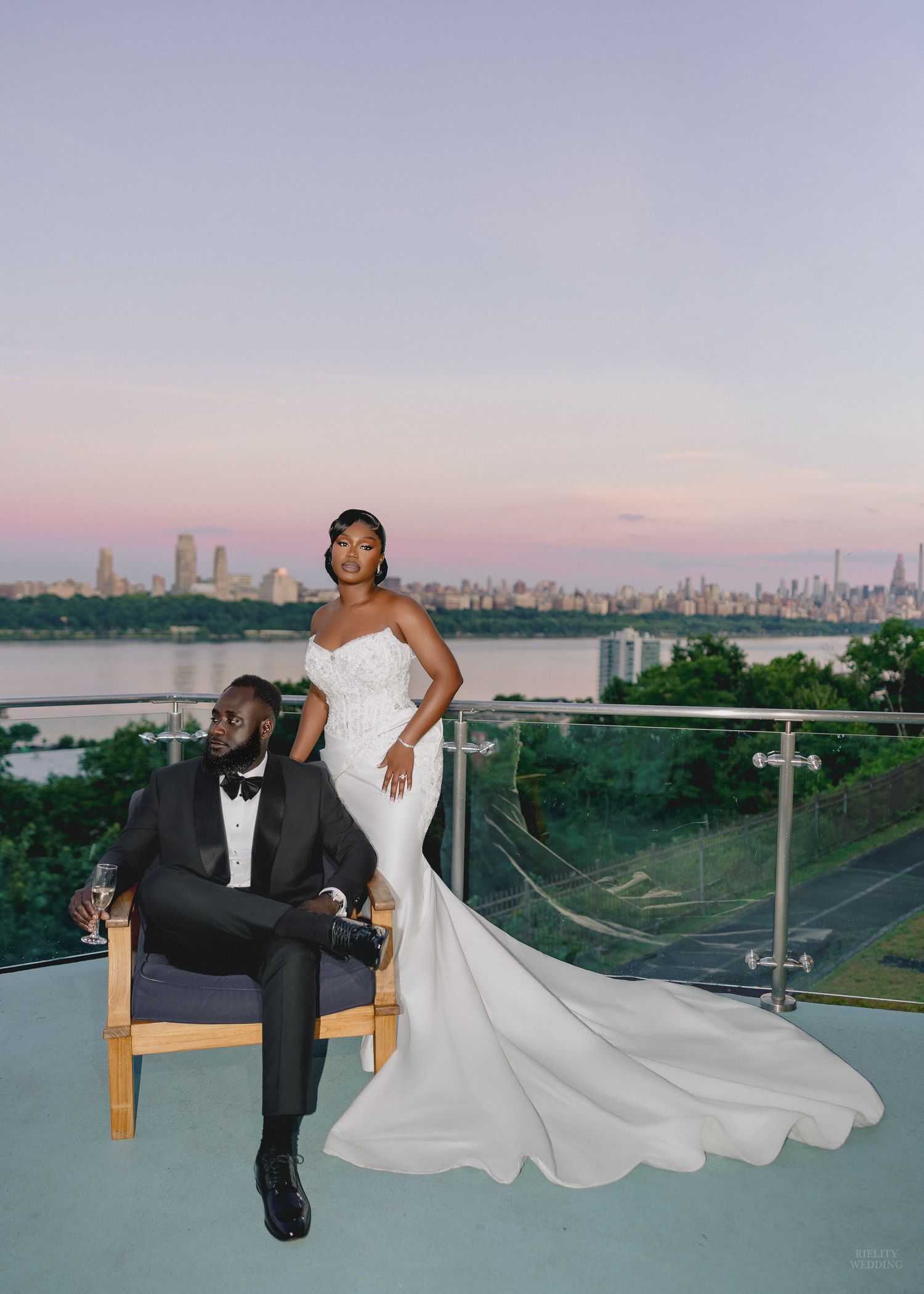 Elegant wedding couple on rooftop at sunset, bride in white mermaid gown, groom in black tuxedo, city skyline backdrop.
