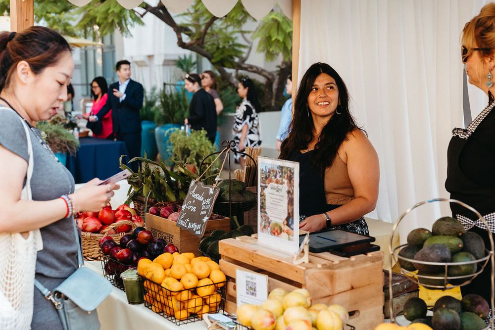 Vendor smiling at a farmers market stall with fresh fruits including lemons and avocados displayed on the table.