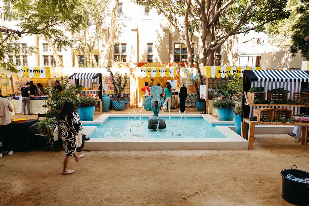Outdoor courtyard with a blue fountain, colorful bunting, market stalls, and lush trees at a vibrant open-air event.