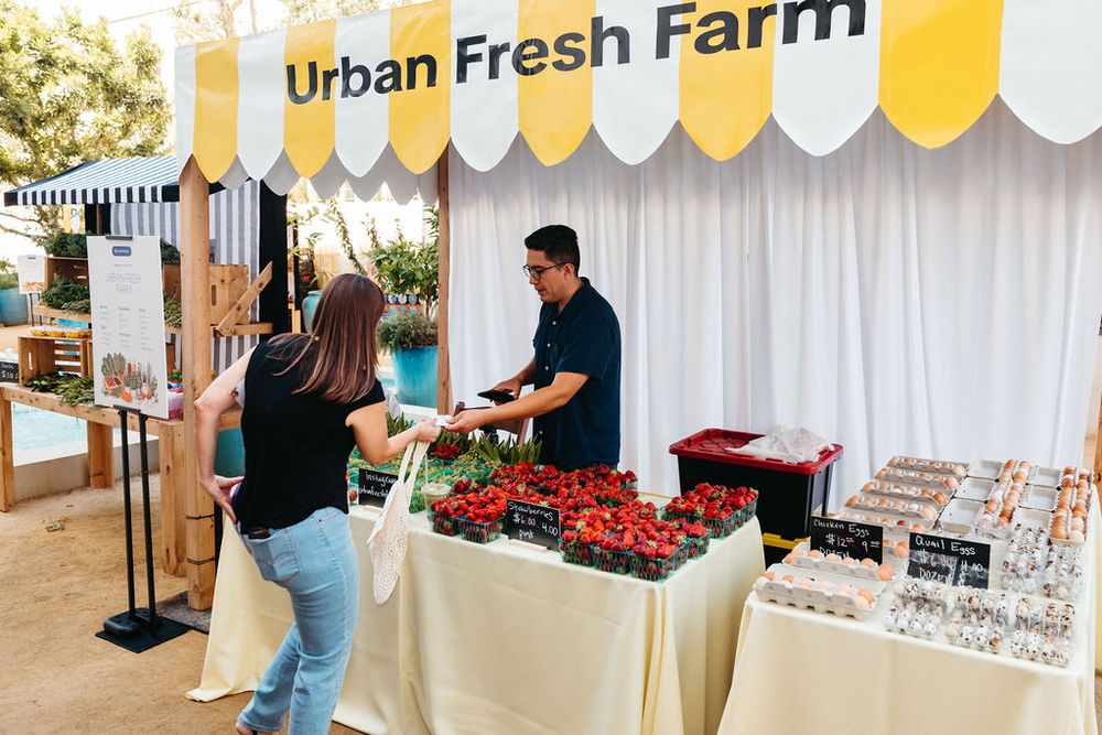 Customer shopping at Urban Fresh Farm market stall with fresh cherries and produce displayed under yellow striped tent.