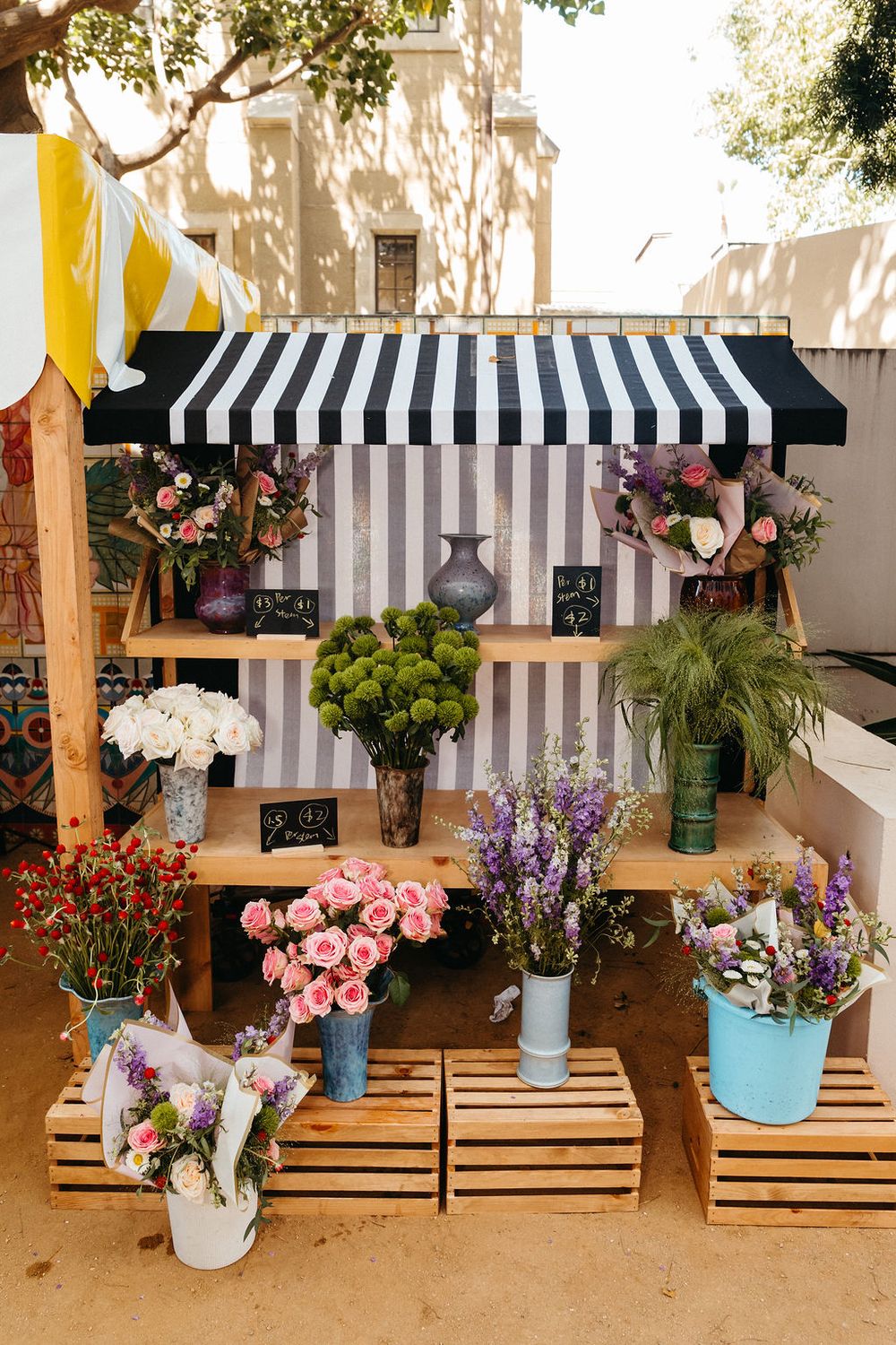 Outdoor flower market stall with wooden shelves displaying colorful bouquets under a black and white striped awning.