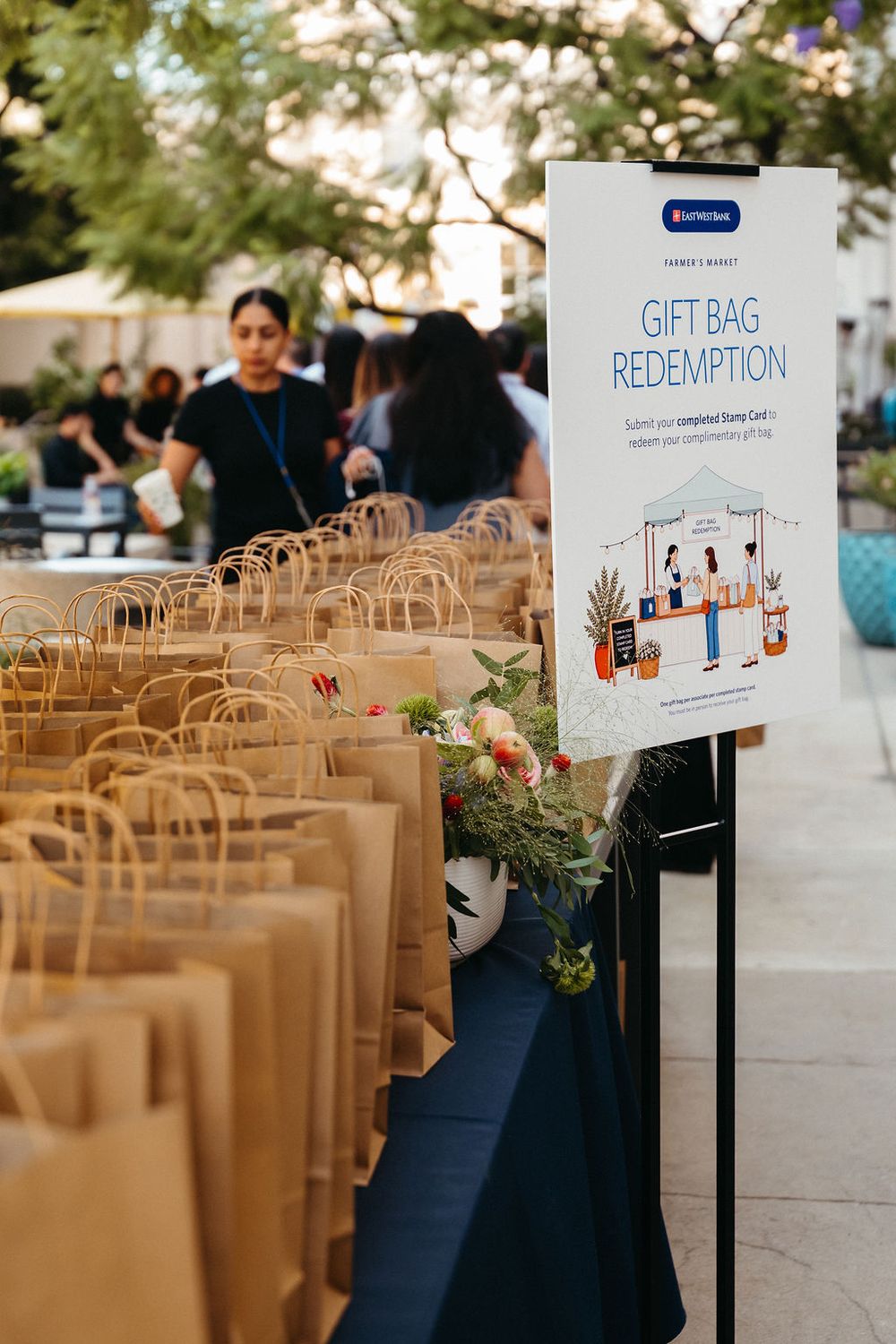 Gift bag redemption station with rows of brown paper bags on a table at an outdoor market event.