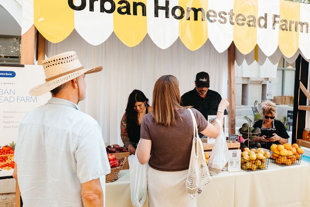 Customers browse fresh produce at the Urban Homestead Farm market stall with yellow and white decor.