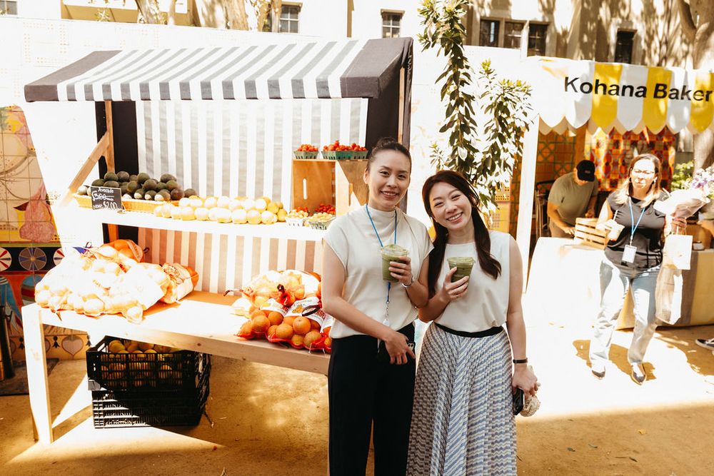 Two women smile and pose together at Kohana Bakery outdoor market stall with fresh fruit displays.