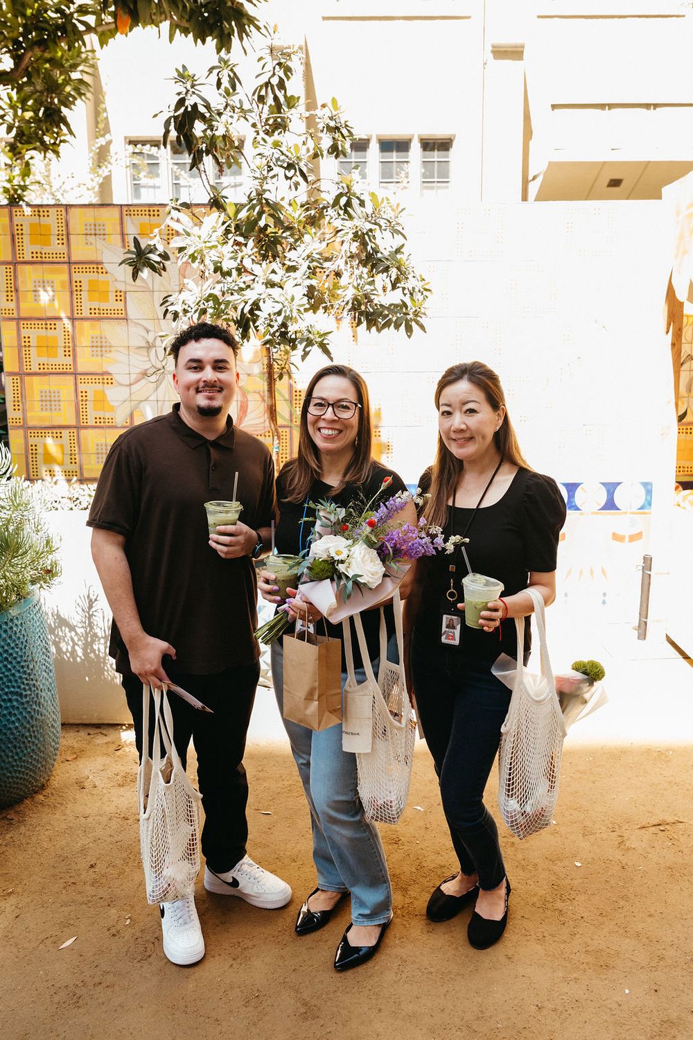 Three people smiling outdoors holding green drinks and flower bouquets in mesh bags at a sunny event.