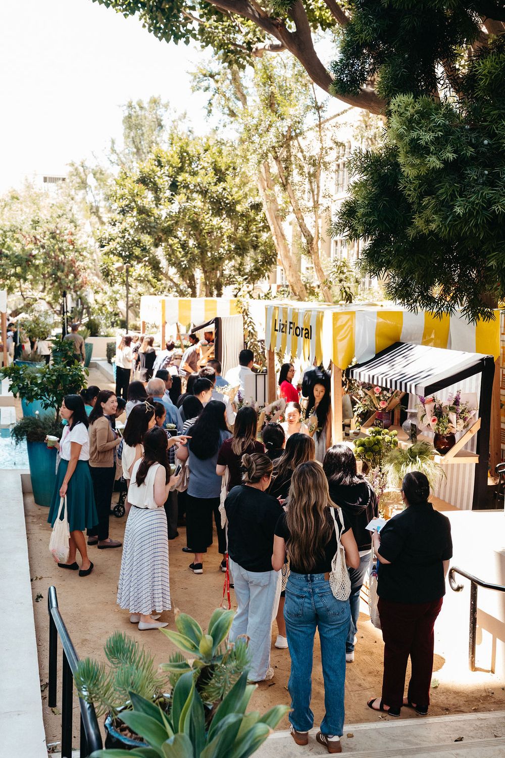 Crowd of people gathered outside at an outdoor market event with yellow vendor booths and lush trees in sunlight.