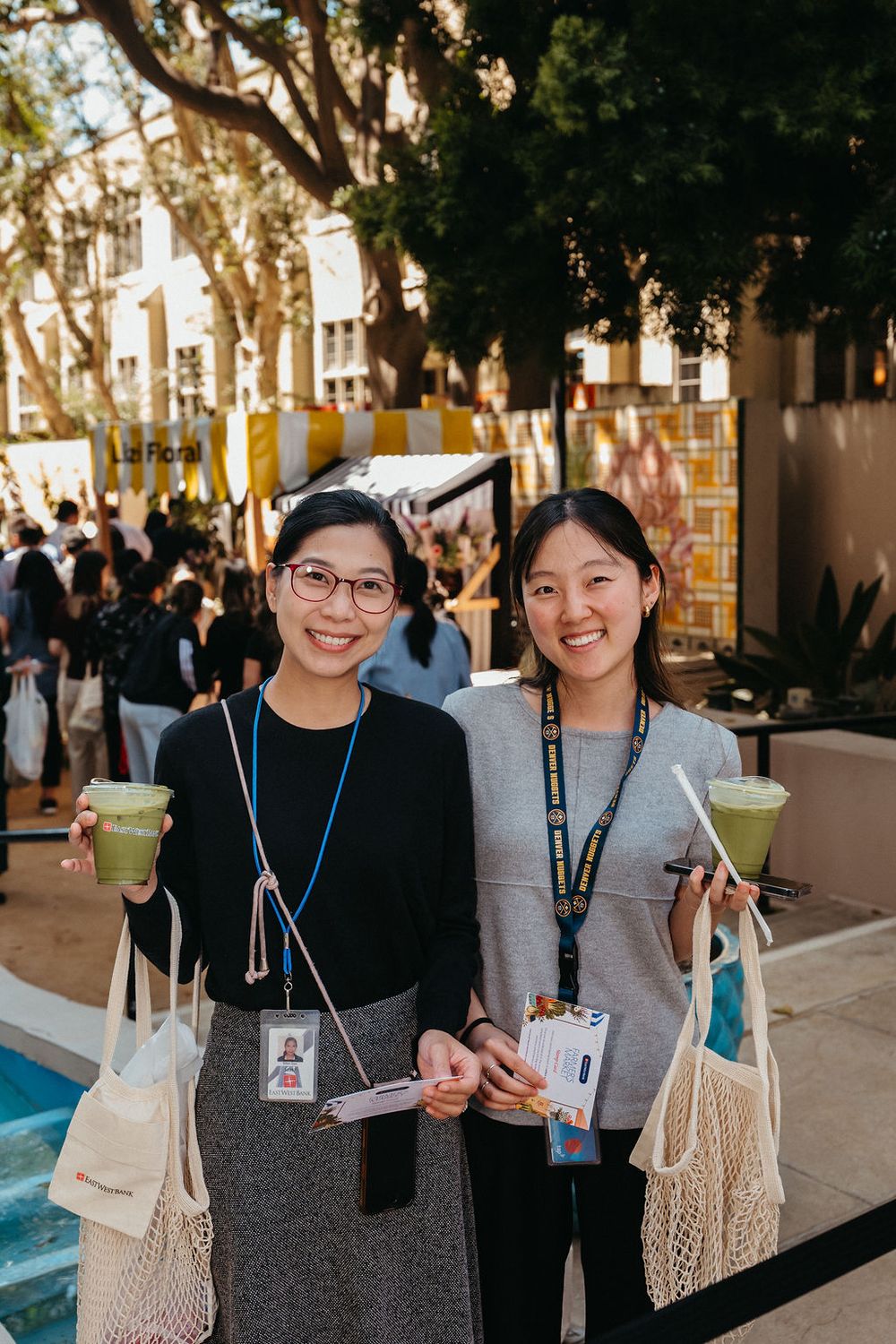 Two smiling women holding green drinks and mesh bags at an outdoor event with trees and yellow signage.